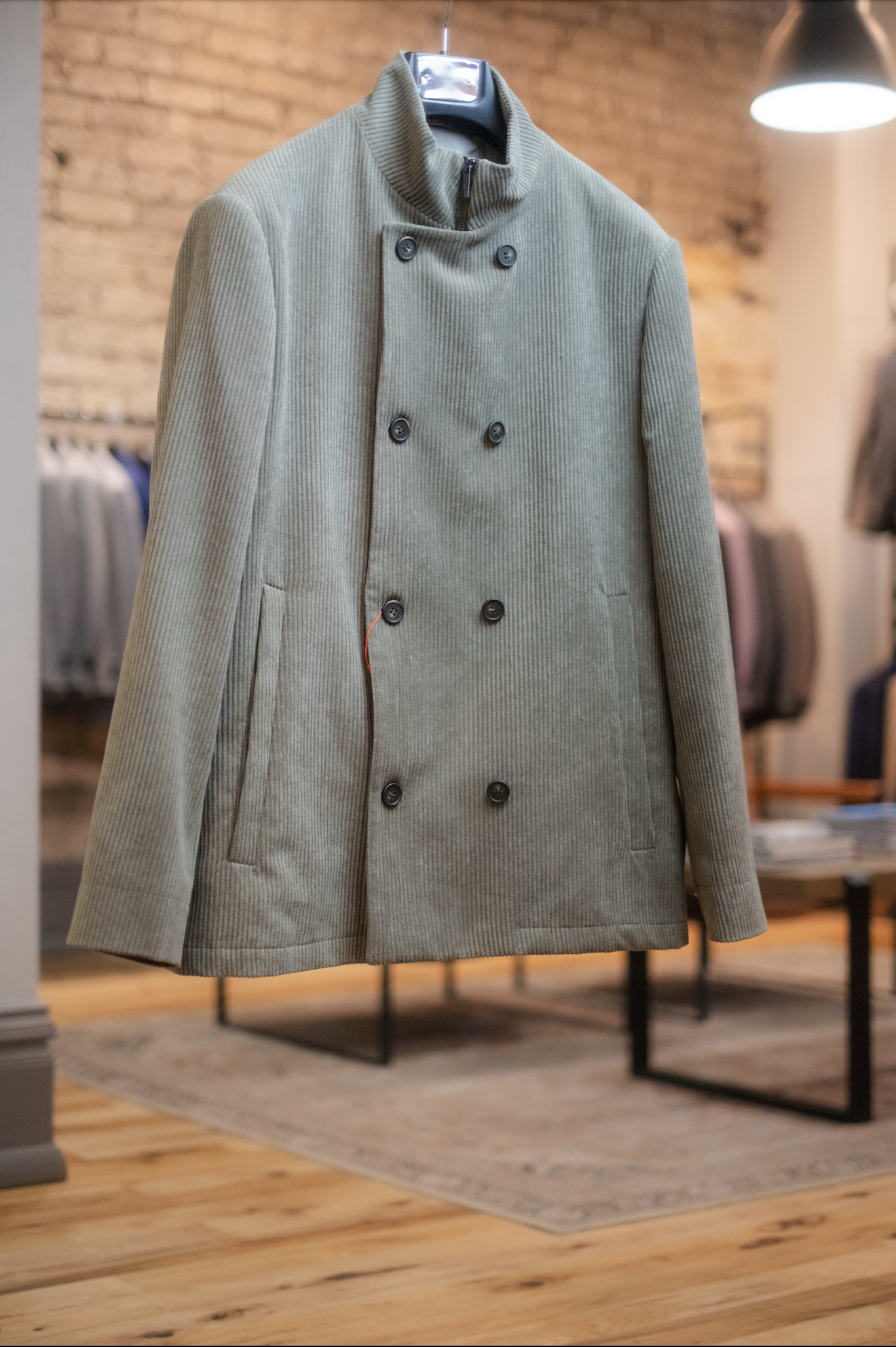 A beige double-breasted coat with a stand-up collar on a hanger inside a clothing store.