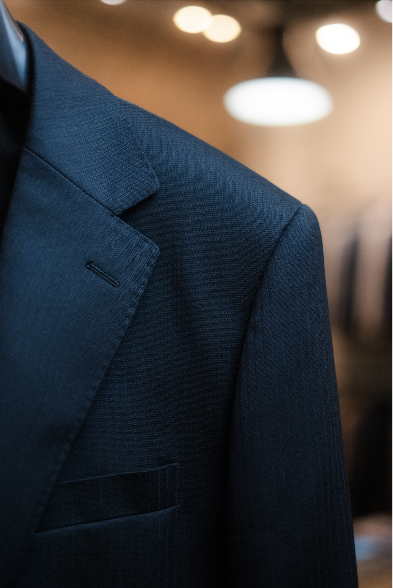 Close-up of a black suit jacket fabric, with out-of-focus ceiling lights in the background.