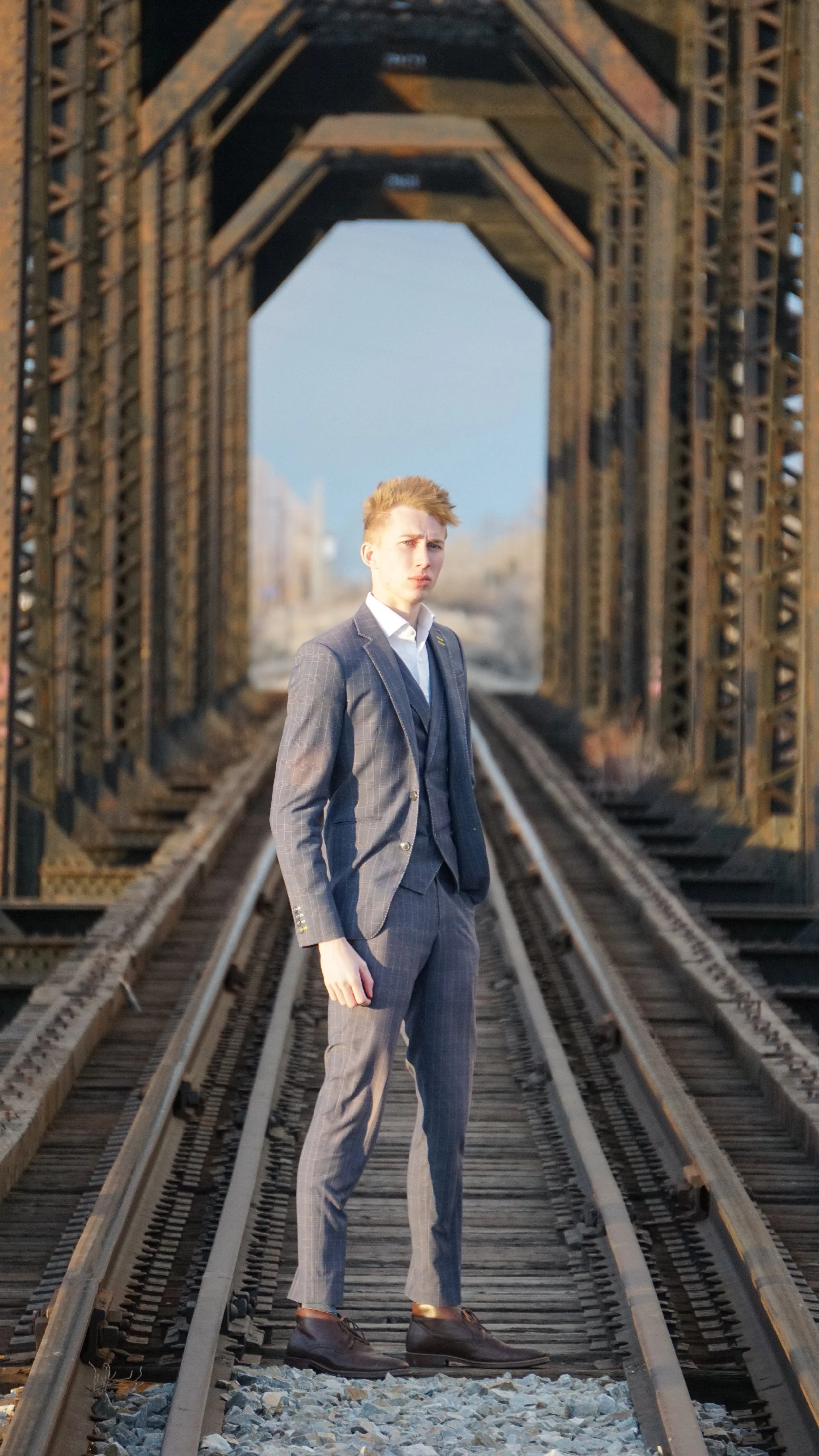 A young man in a gray plaid suit standing on train tracks under a metal bridge, with trees and sky in the background.