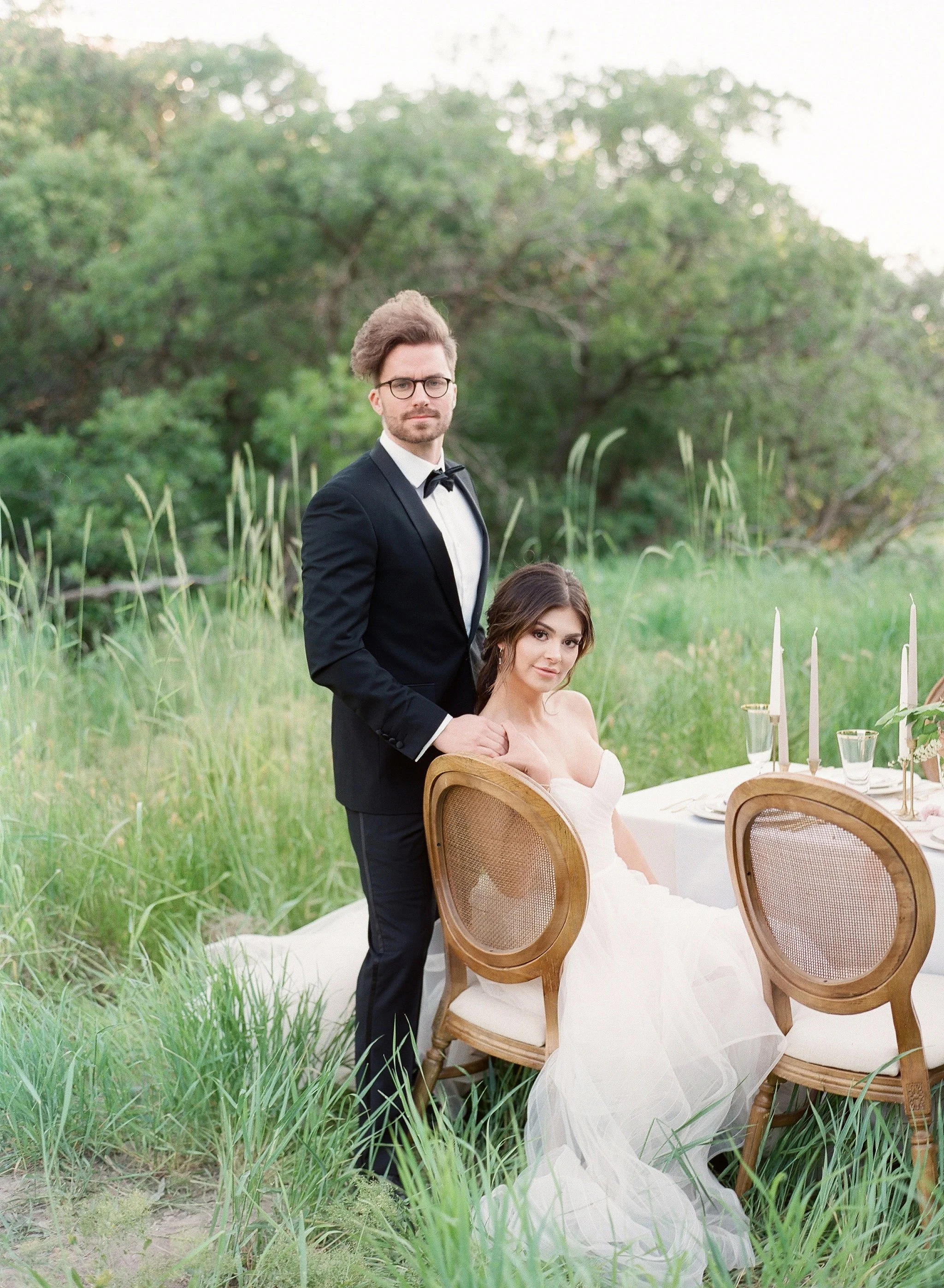 A man in a tuxedo with glasses standing next to a seated woman in a white gown at an outdoor event with green trees and tall grass in the background.