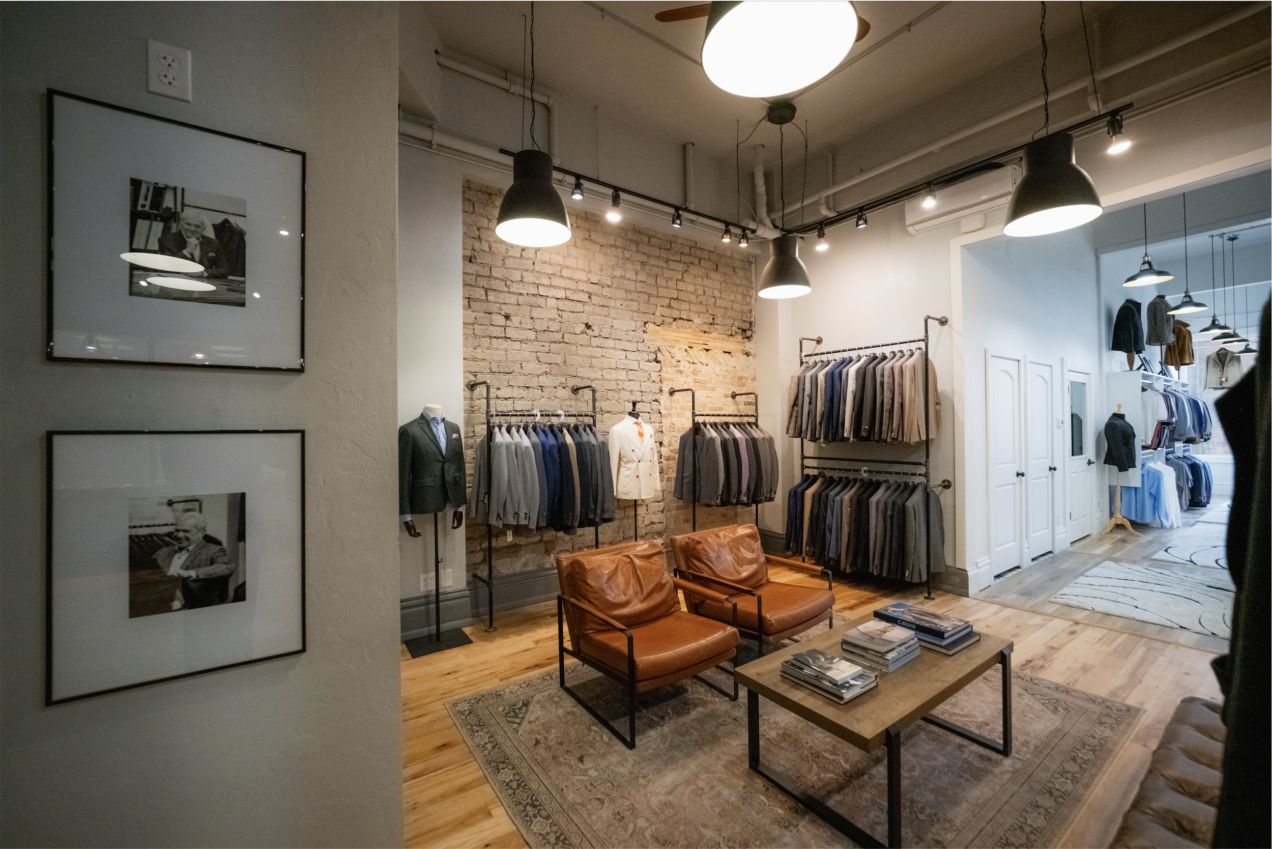 Interior of a clothing store with hanging lights, a brick accent wall, and racks of shirts and jackets. There are chairs and a coffee table with books in the foreground, and framed black-and-white photographs on the wall to the left.