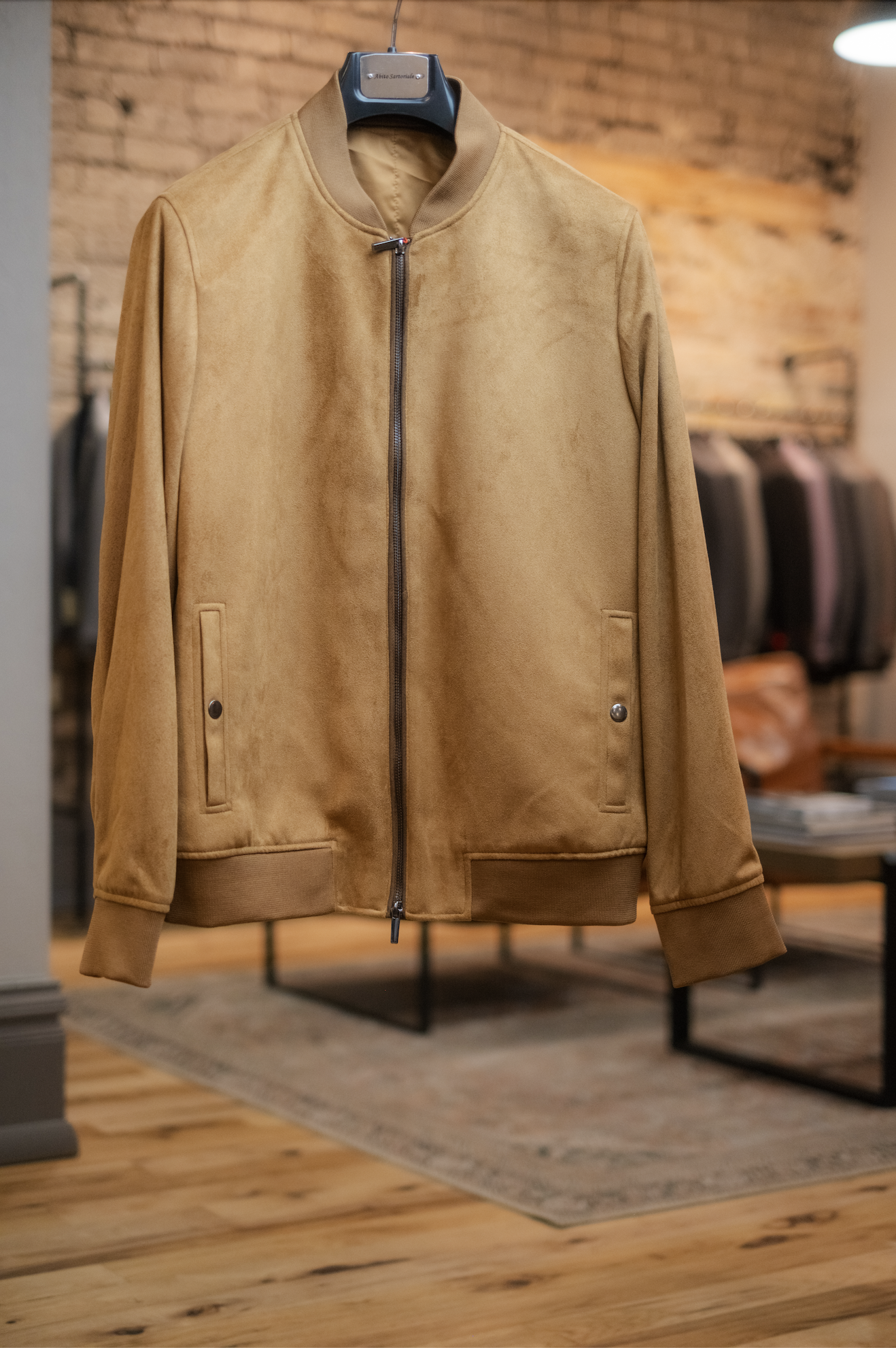 A tan suede bomber jacket hangs on a black hanger in a clothing store.