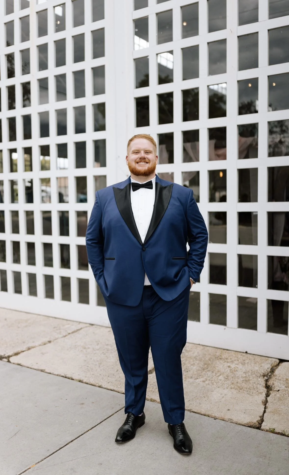 A man in a blue tuxedo with black lapels and a black bowtie standing outdoors in front of a white lattice wall, smiling with his hands in his pockets.