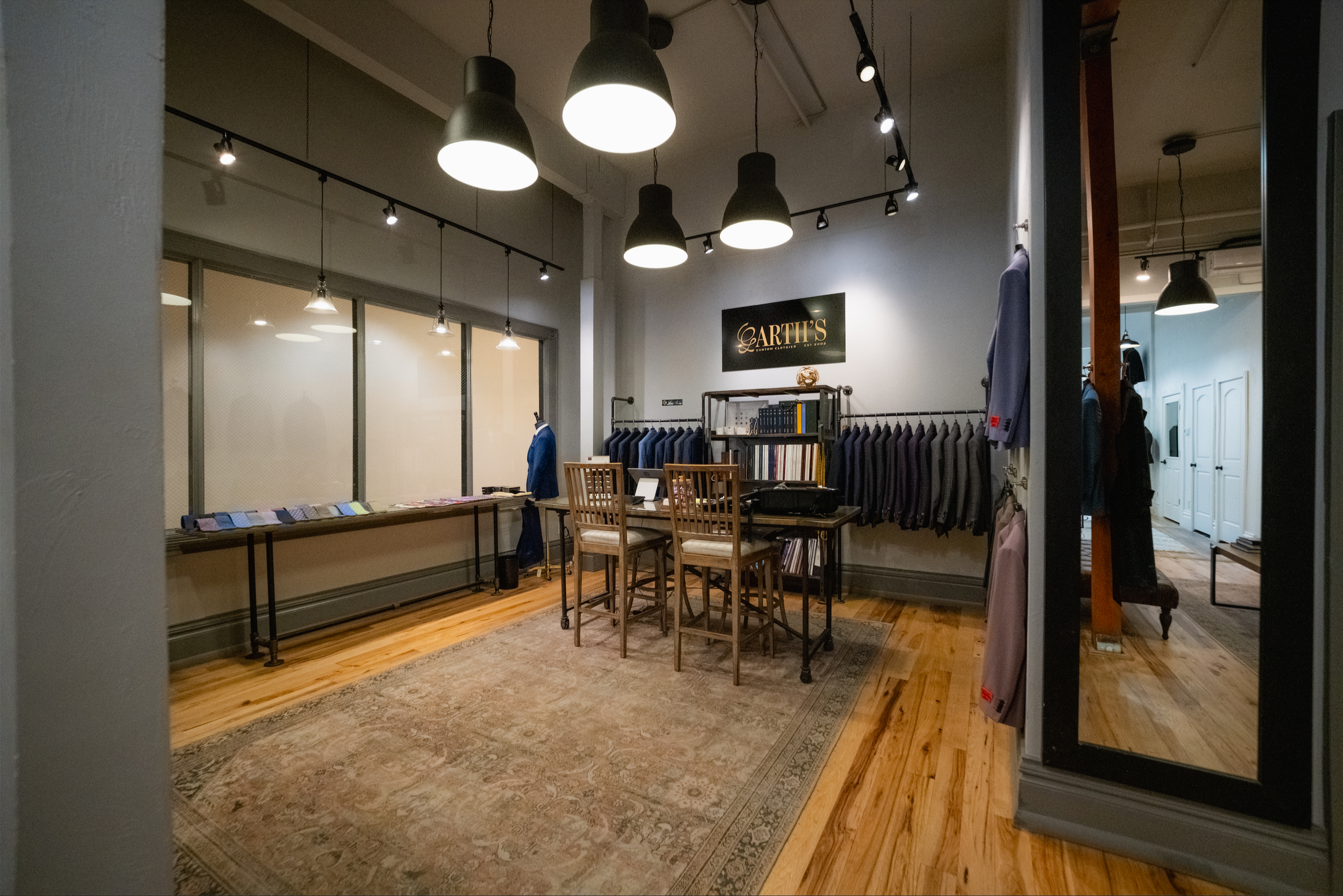 Interior of a clothing store with hanging lights, racks of suits, and a table with fabric swatches.