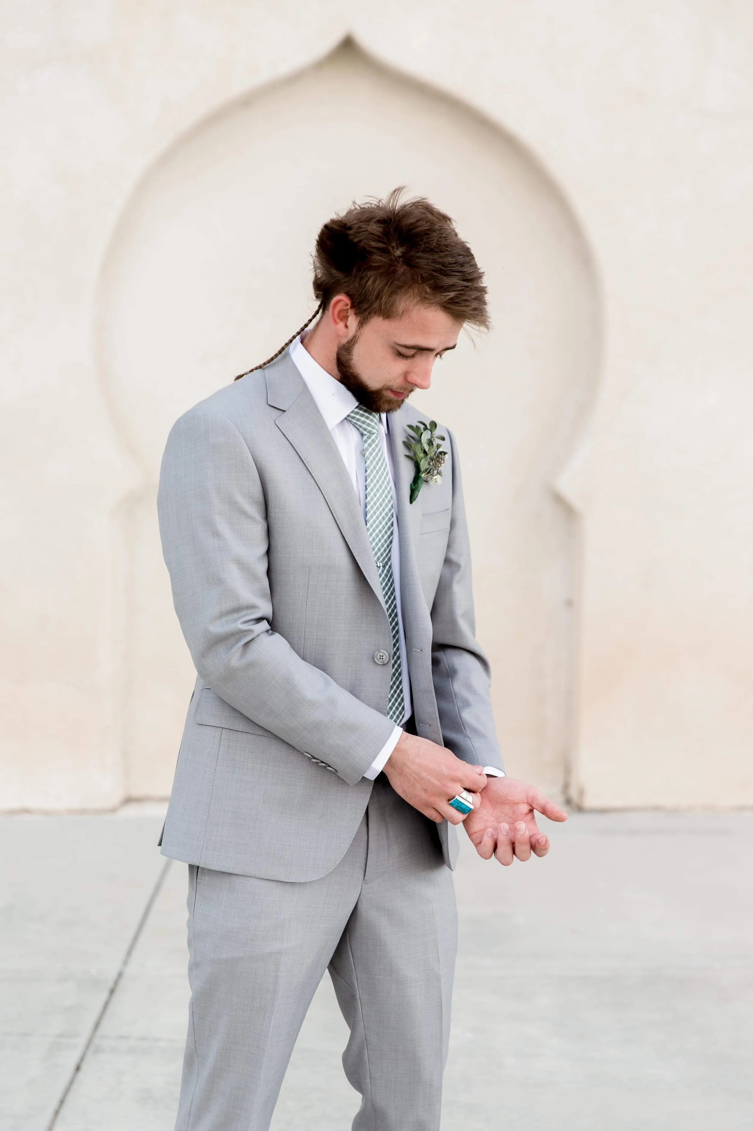 A man in a light gray suit with a boutonniere, looking down and adjusting his sleeve in front of an off-white wall with a decorative arch.