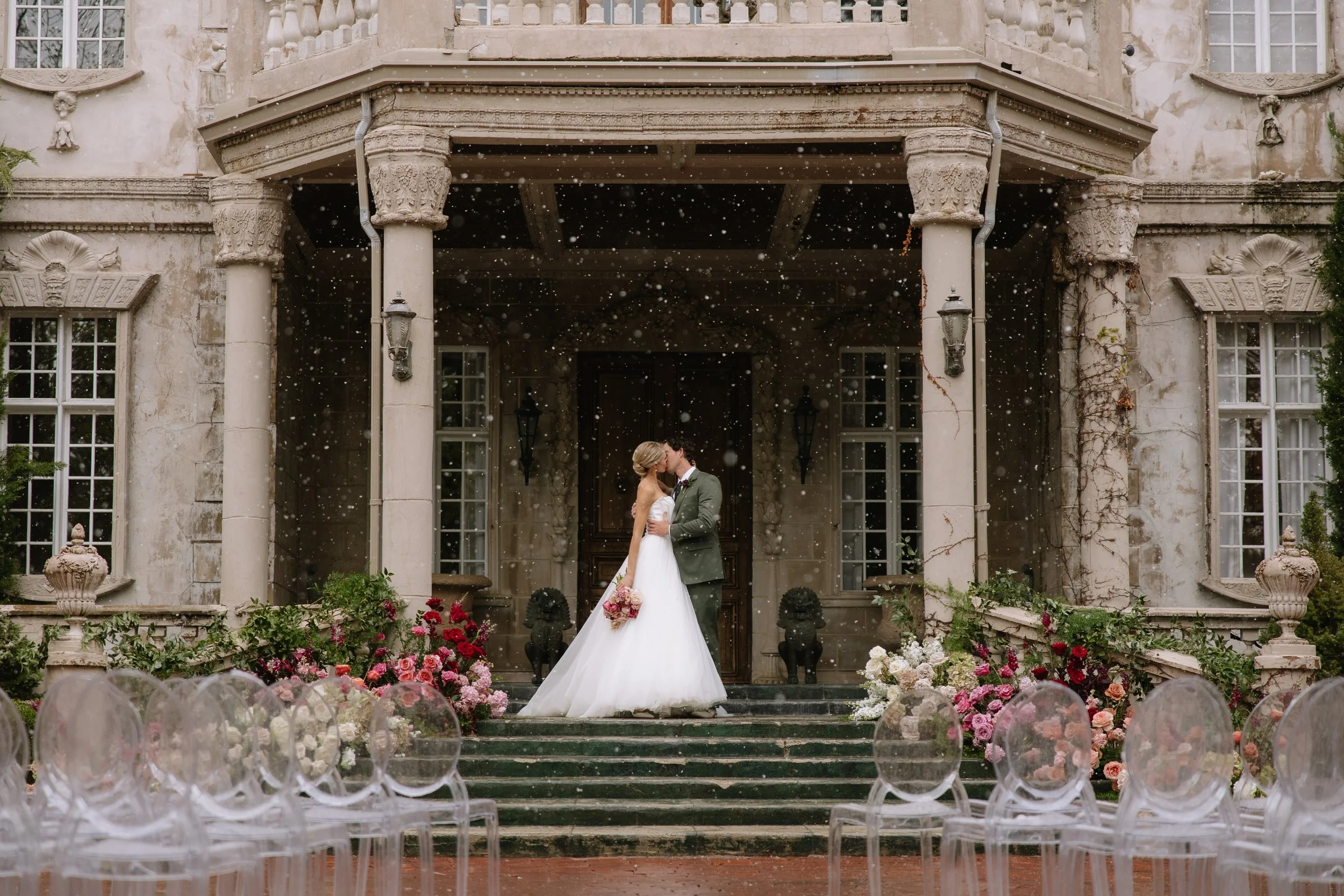 A bride and groom sharing a kiss on the steps of a vintage mansion, with rows of clear chairs and colorful flowers arranged around the area.