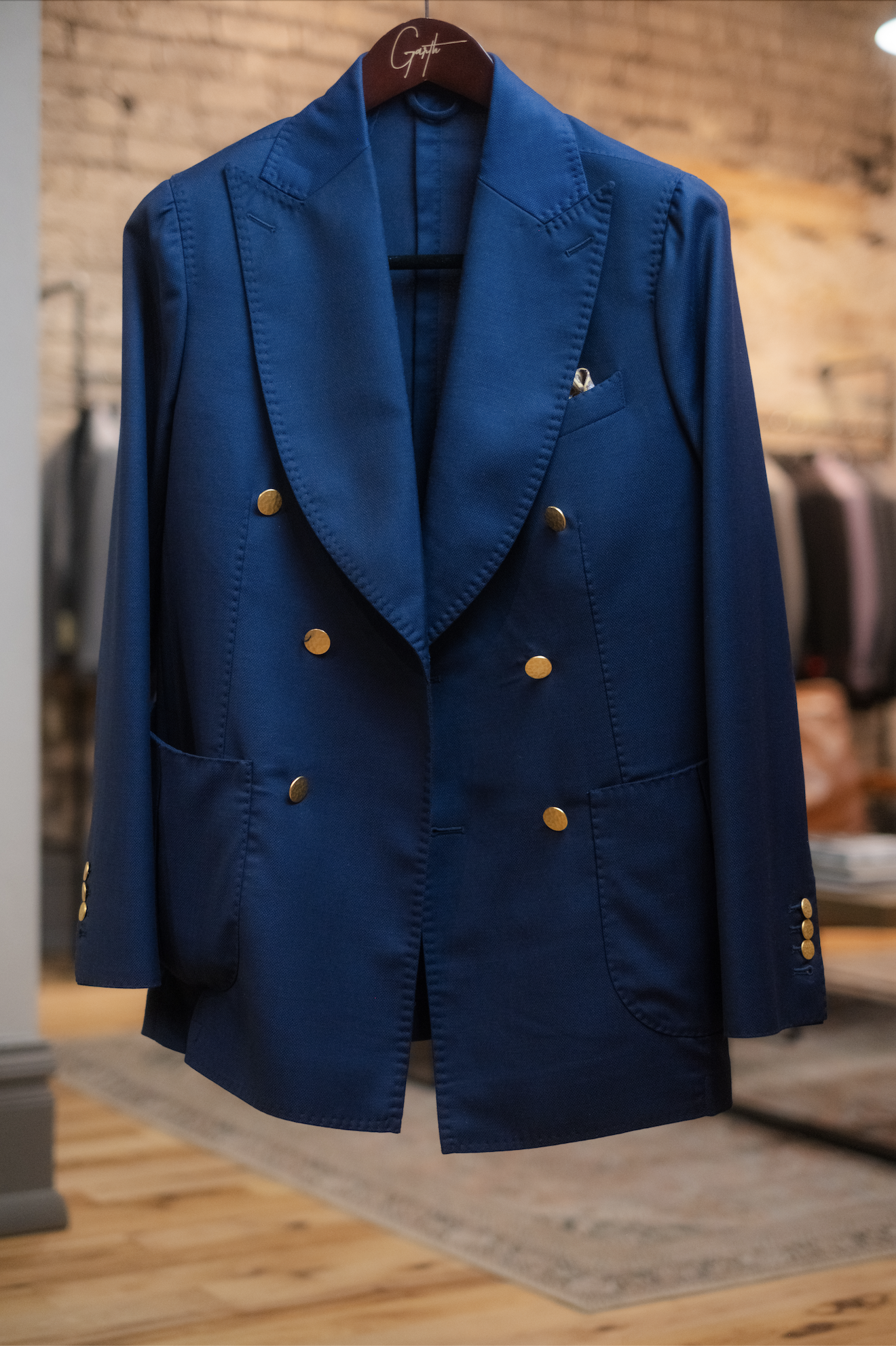 A blue blazer with gold buttons hanging on a wooden hanger in a clothing store, with other clothes visible in the background.