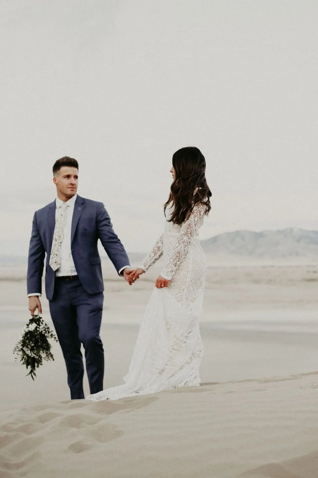 A bride and groom holding hands on a sandy beach, with the groom holding a bouquet of greenery, during their wedding photoshoot.