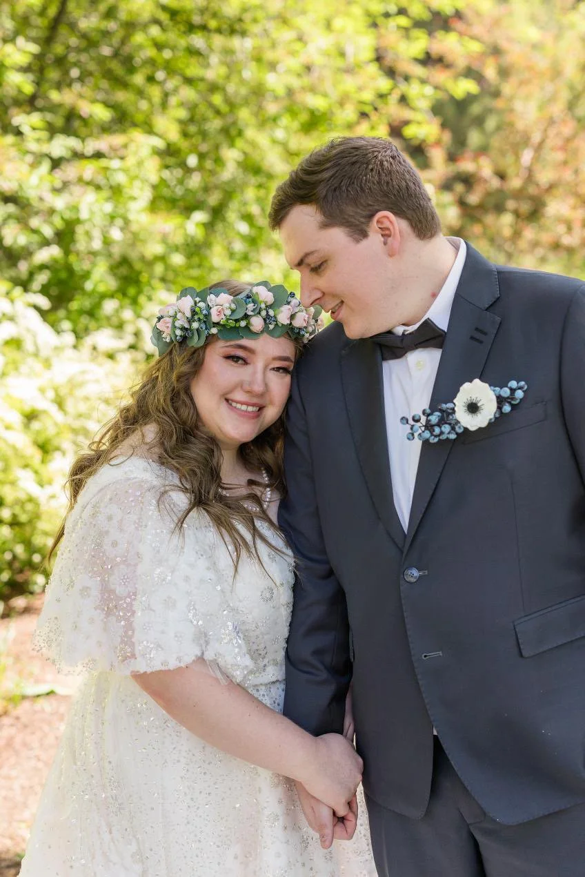 A smiling bride and groom holding hands outdoors, with the groom leaning his head on the bride's head. The bride is wearing a white wedding dress and a floral crown, while the groom is dressed in a dark suit with a white shirt and black bow tie, and 