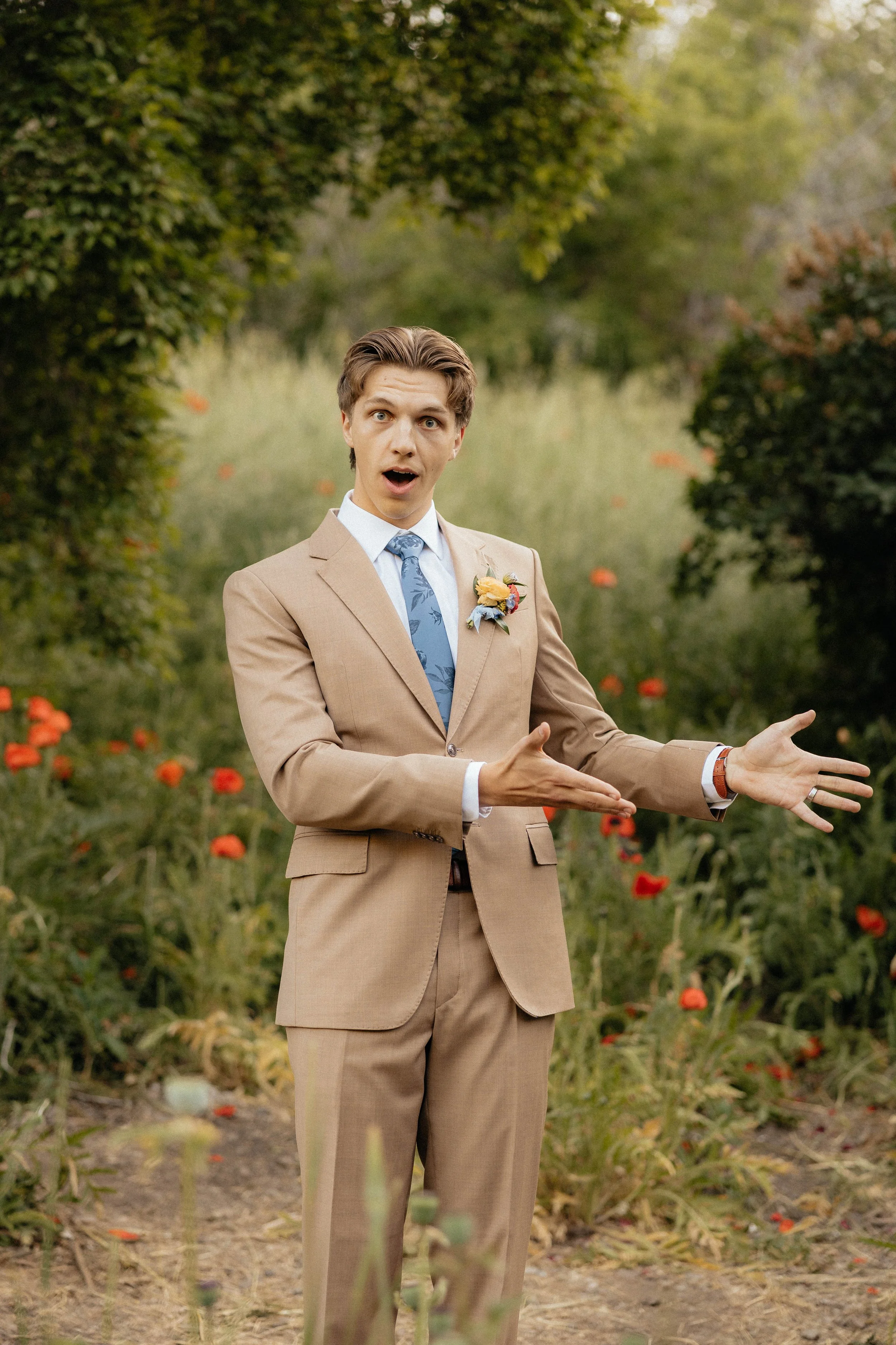 A man dressed in a tan suit with a floral tie and boutonniere, standing outdoors in a garden with green trees and red flowers, gesturing with his hands and surprised expression.