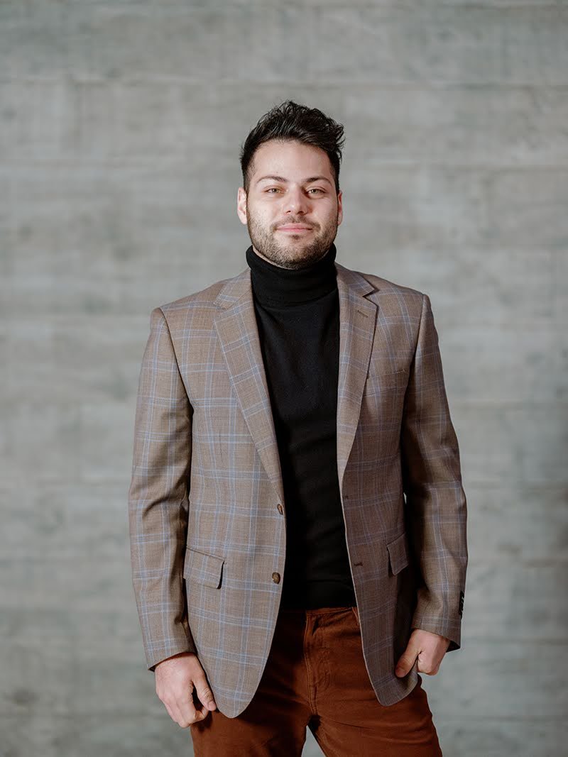 A man with dark hair, styled upwards, wearing a plaid blazer over a black turtleneck, standing against a light gray wooden wall background.