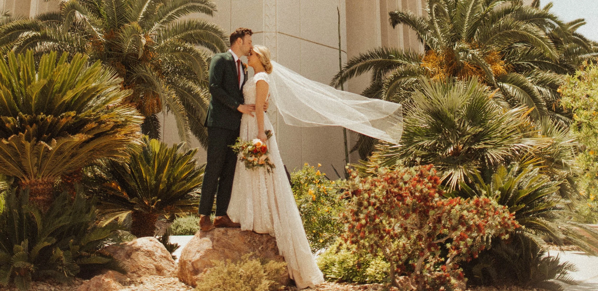 A bride and groom sharing a kiss outdoors surrounded by lush green plants and colorful flowers during their wedding ceremony.