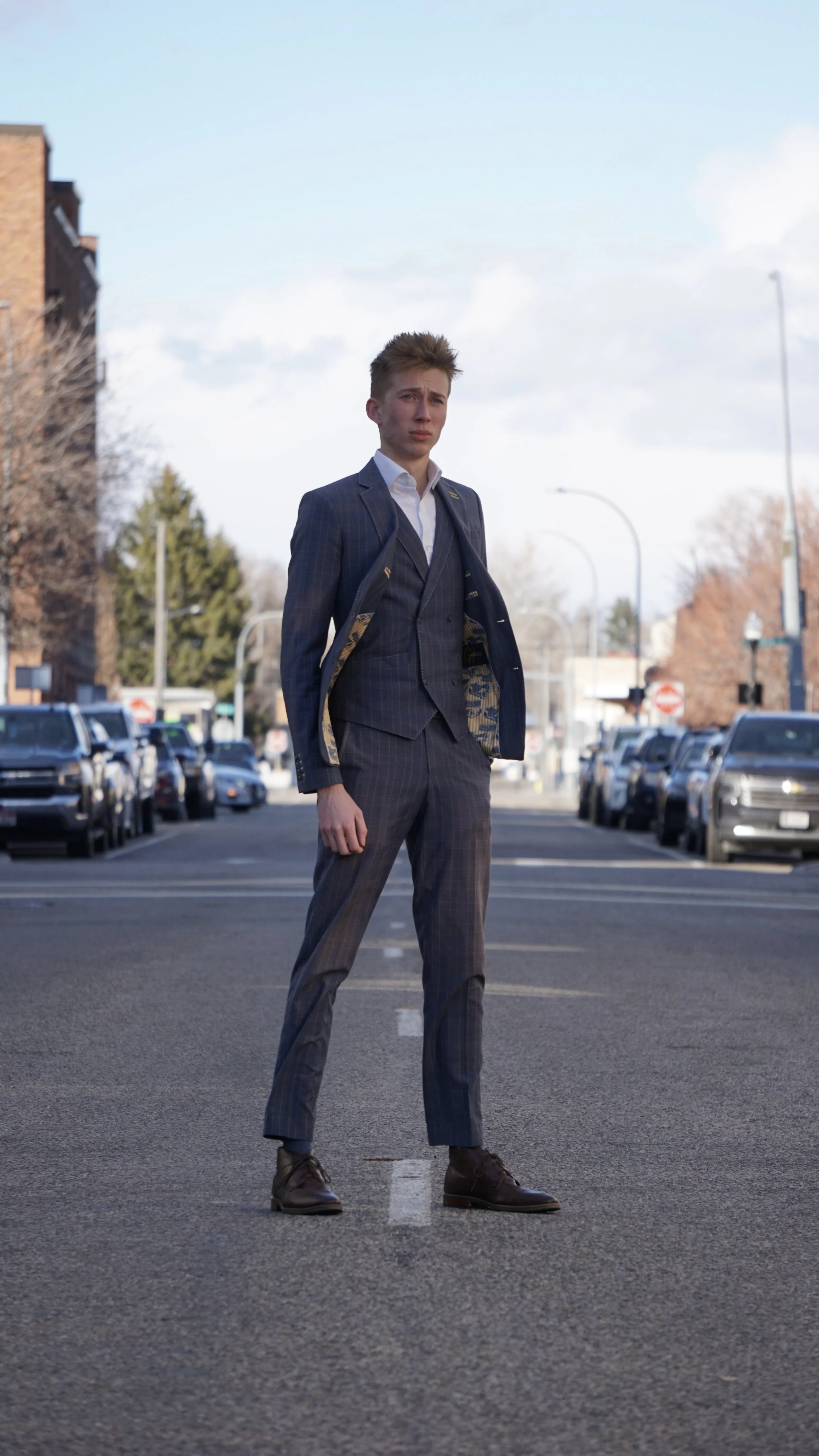 Young man in a suit standing in the middle of a street, with cars parked on both sides and trees and buildings in the background.