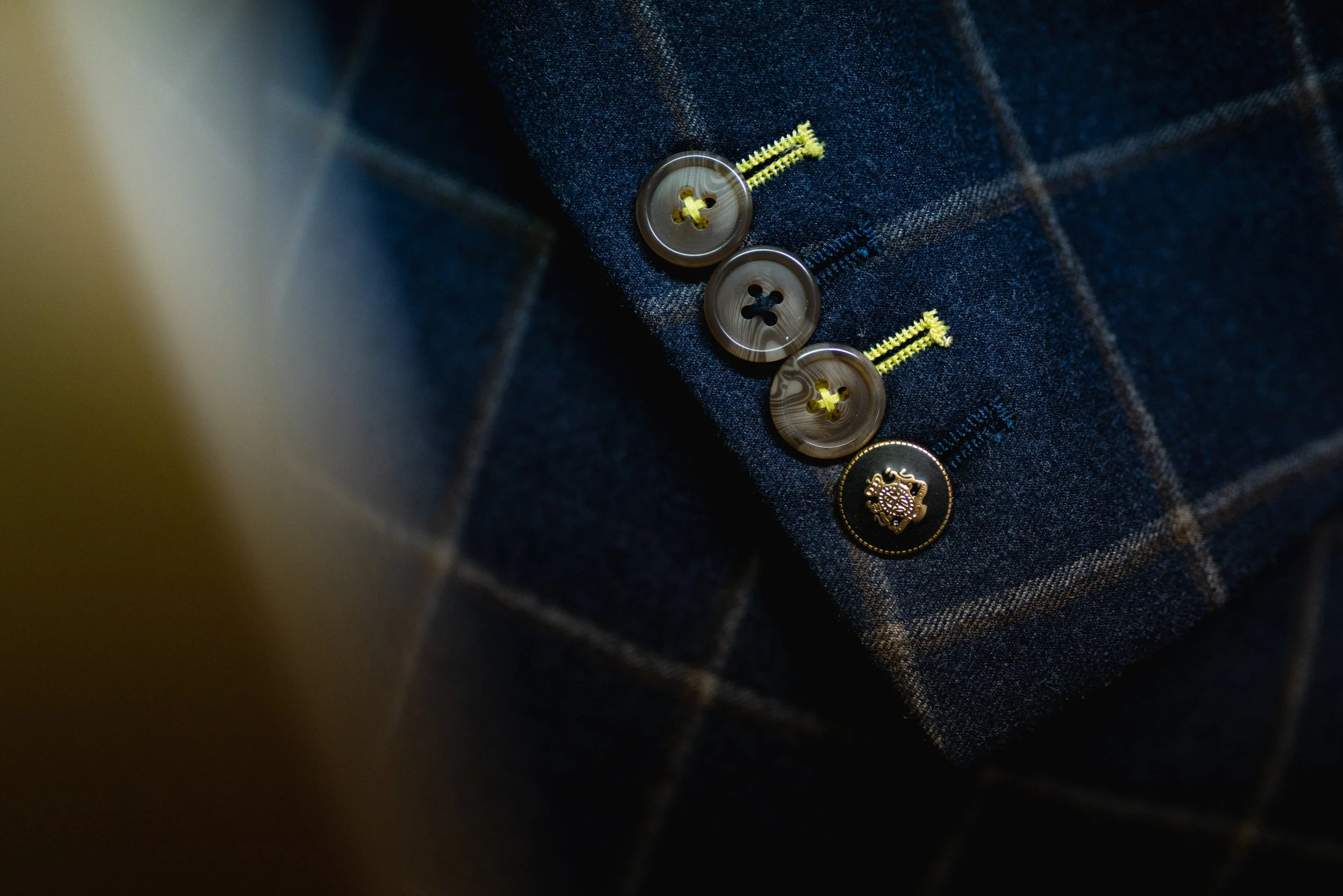 Close-up of four buttons sewn onto a dark windowpane fabric, with yellow stitching and a unique button with a lion crest.