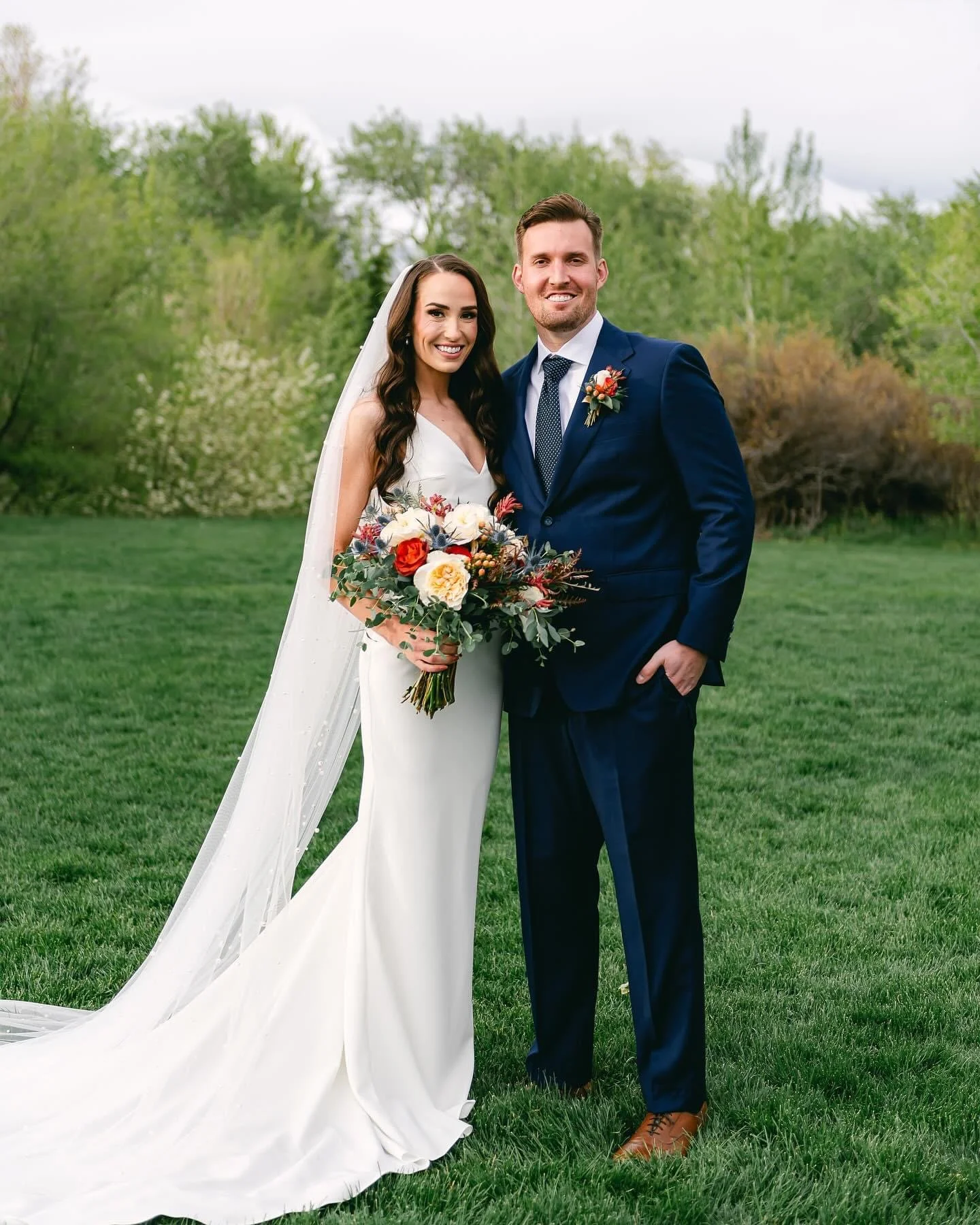 A bride and groom standing outdoors on grass, smiling, with trees and cloudy sky in the background. The bride wears a white wedding gown with a veil and holds a bouquet of white, red, and purple flowers. The groom wears a navy blue suit, white shirt,