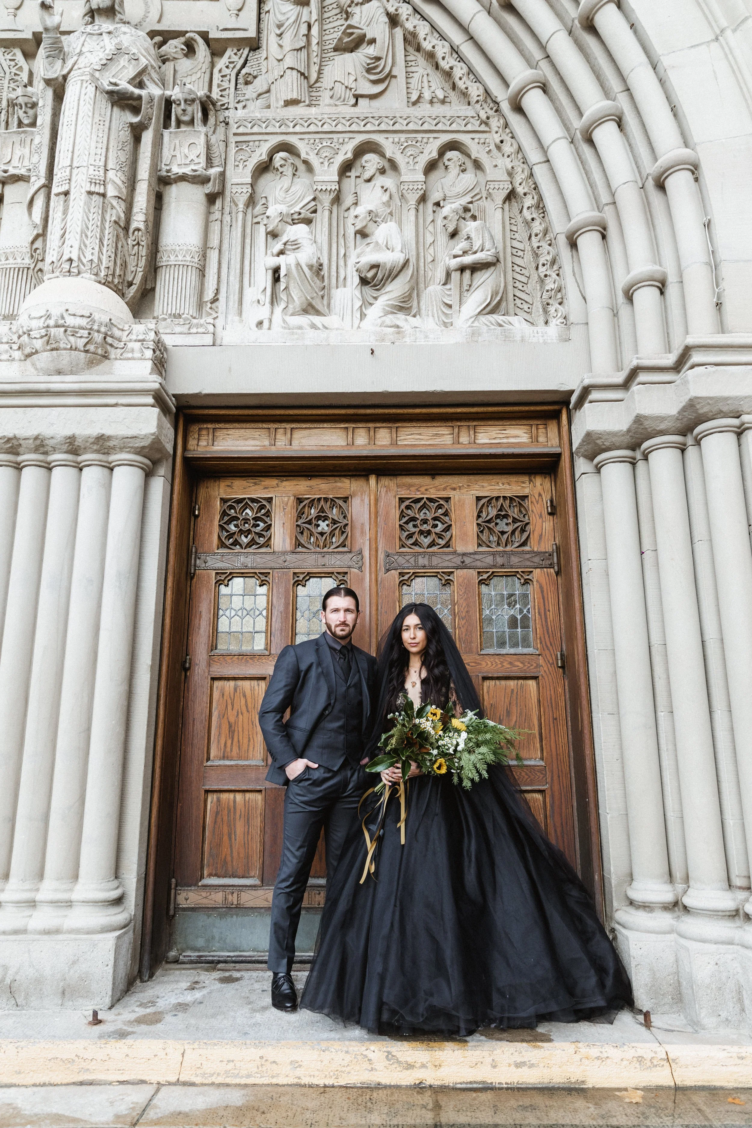 A couple stands in front of a carved stone church entrance, with the woman holding a bouquet of flowers and wearing a long black dress and veil, and the man dressed in a dark suit with his hand in his pocket.
