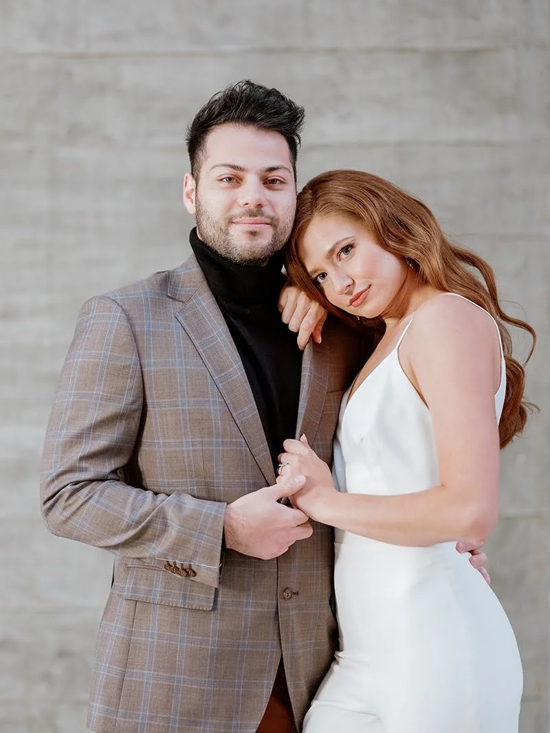 A couple standing closely together, posing for the camera against a neutral background. The man has dark hair, a beard, and is dressed in a plaid blazer and black turtleneck. The woman has long red hair and is wearing a white sleeveless dress. They b