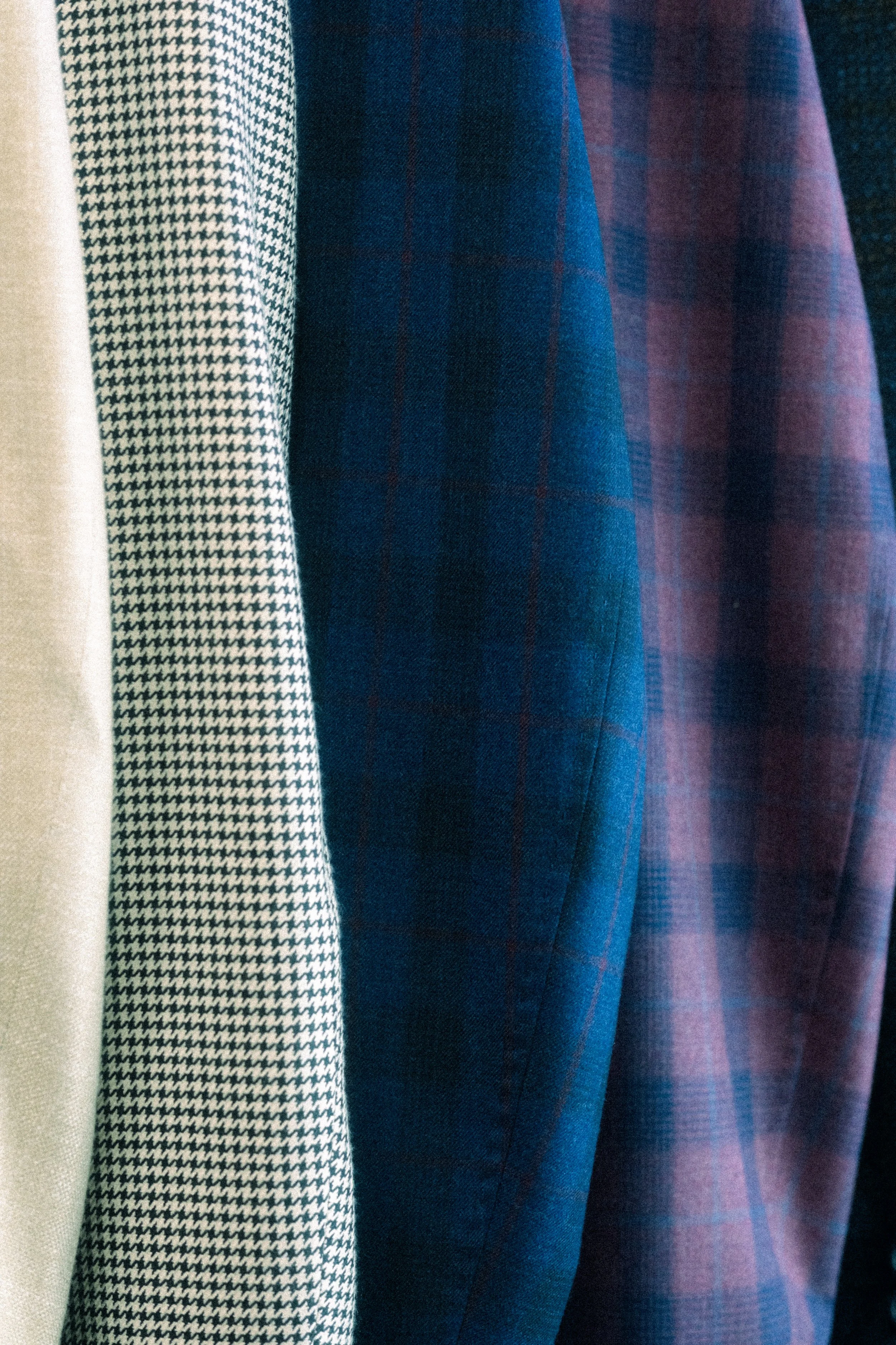 Close-up of various fabric patterns and colors hanging, including houndstooth, plain beige, dark blue with red plaid, and purple plaid.