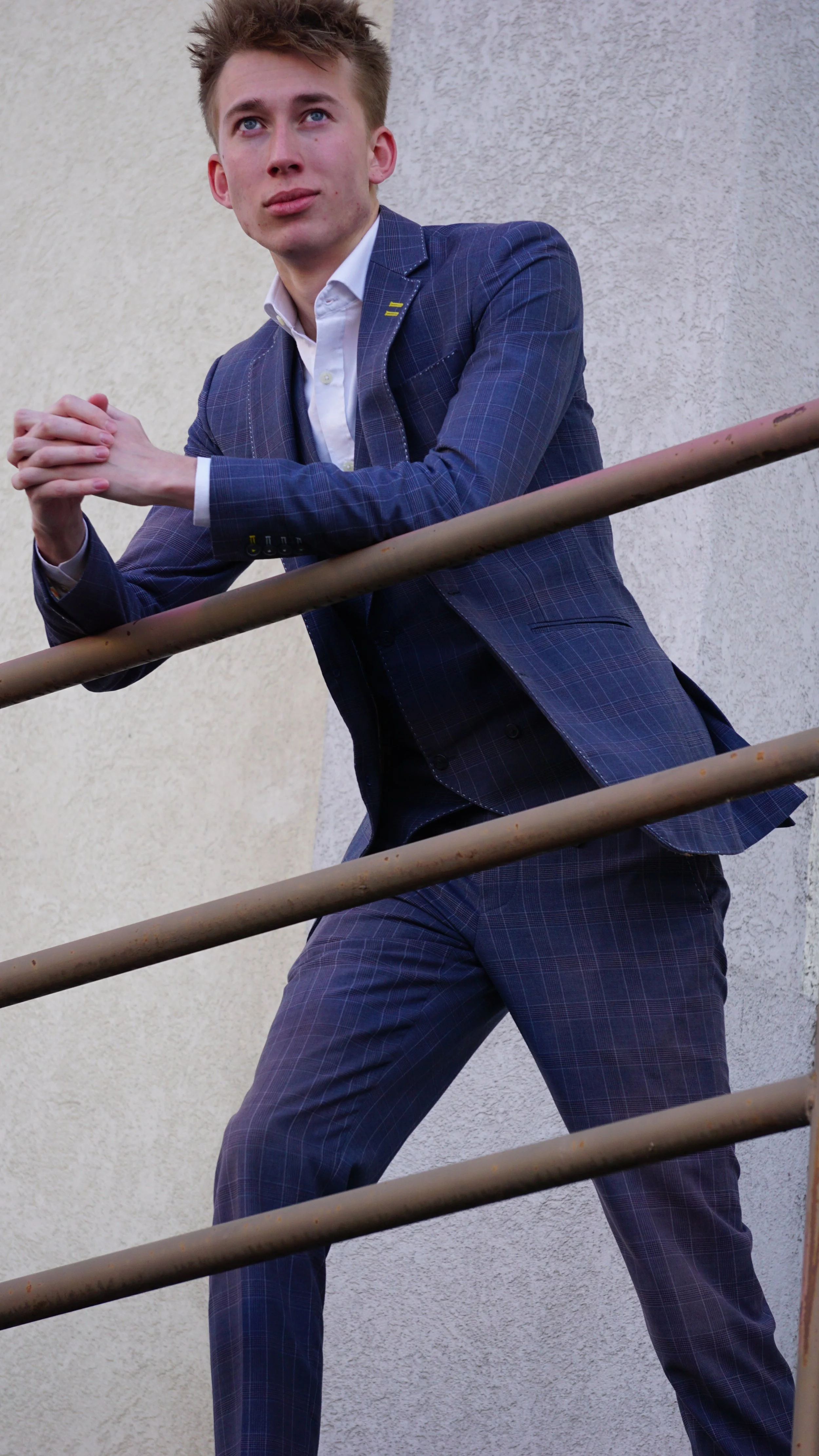 A young man in a blue plaid suit with a white dress shirt leaning against a beige metal railing outdoors, with a white textured wall behind him.