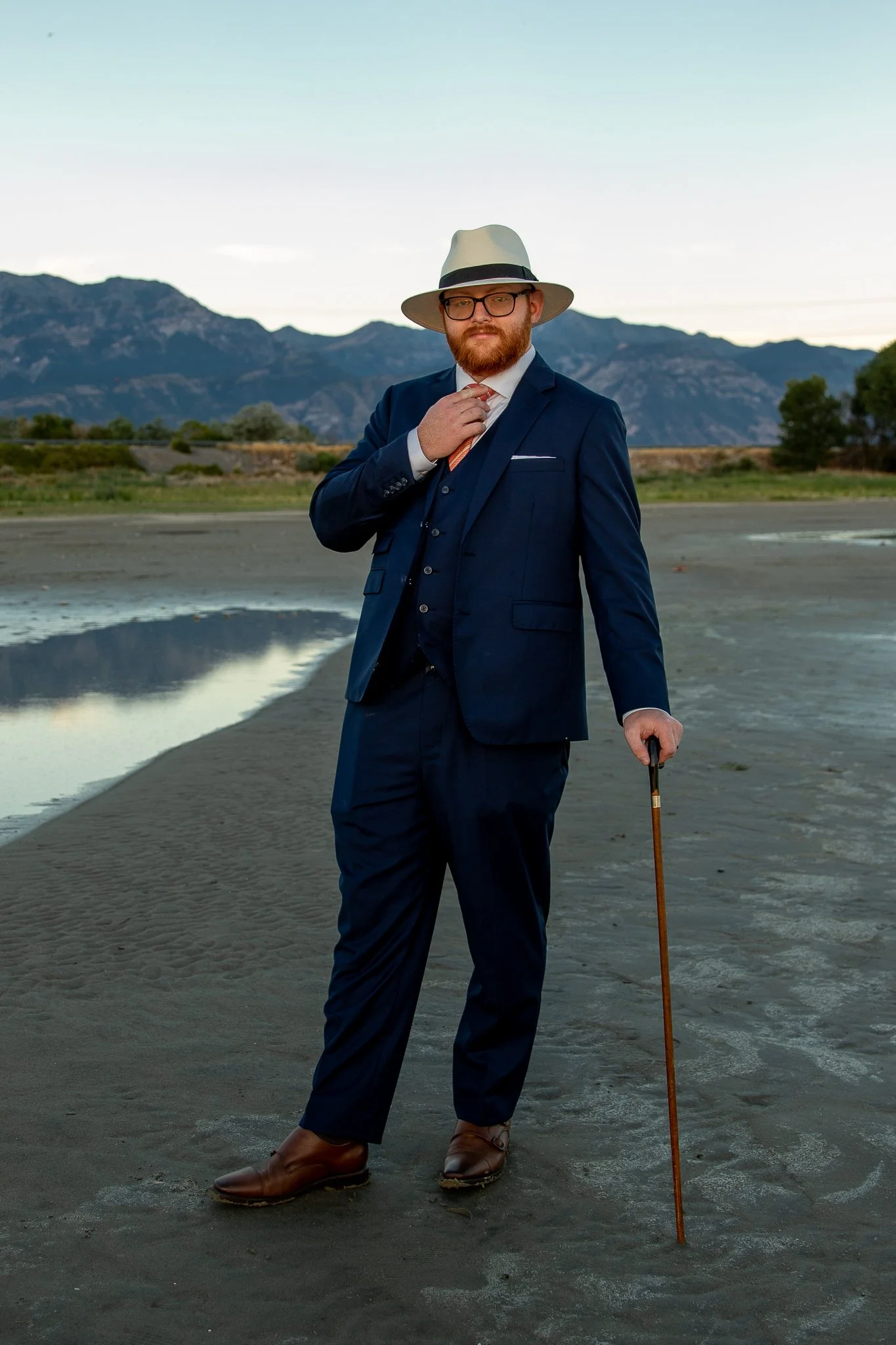 A man wearing a navy blue suit, white shirt, orange tie, brown shoes, and a white fedora with a black band stands outdoors on a sandy beach or dried riverbed with mountains in the background. He is holding a walking cane and adjusting his tie, with a