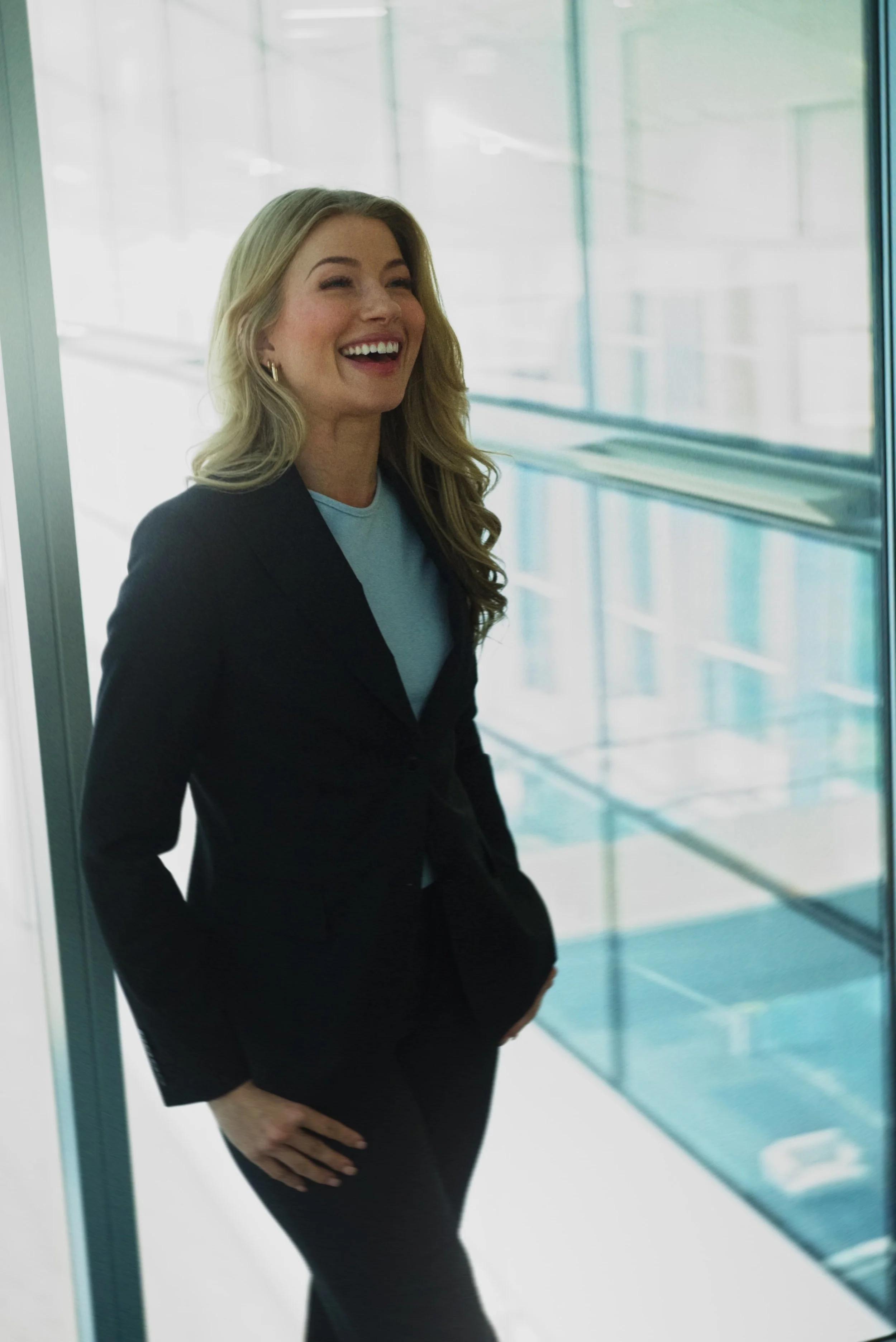 A woman with blonde hair smiling and wearing a black blazer and black pants, standing indoors by a glass wall with reflections of buildings outside.