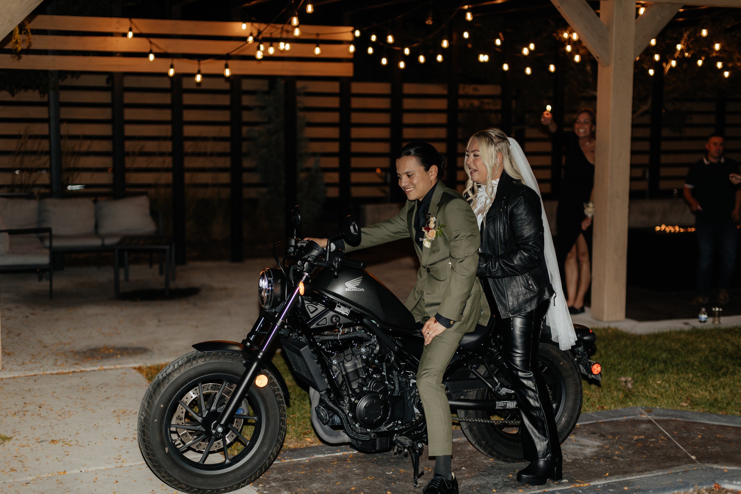 A person in a green suit riding a black Harley-Davidson motorcycle with a woman in black leather jacket sitting behind them during a nighttime outdoor gathering under string lights.