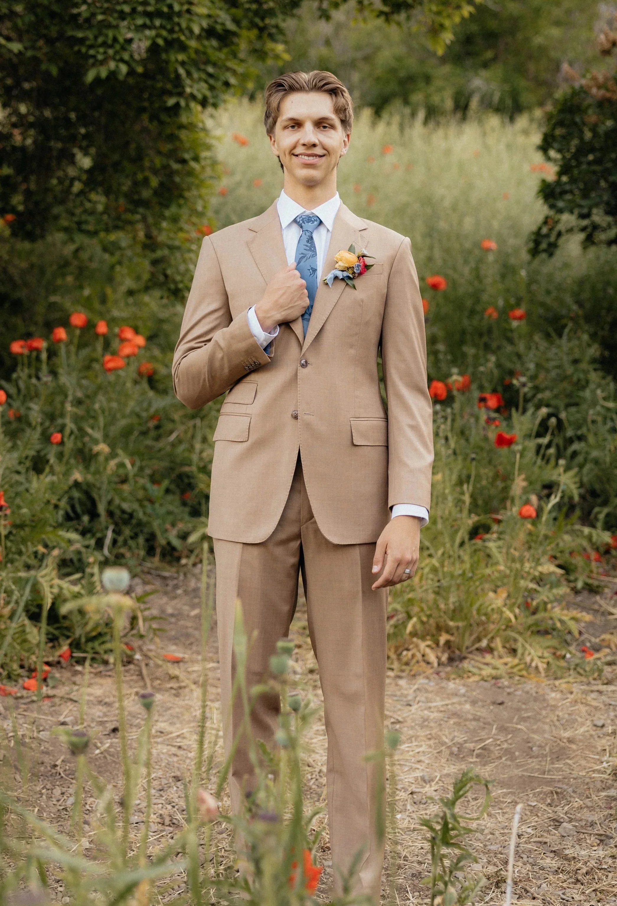 A young man in a tan suit and blue tie, standing outdoors in a garden with red flowers, smiling and holding his lapel with one hand.