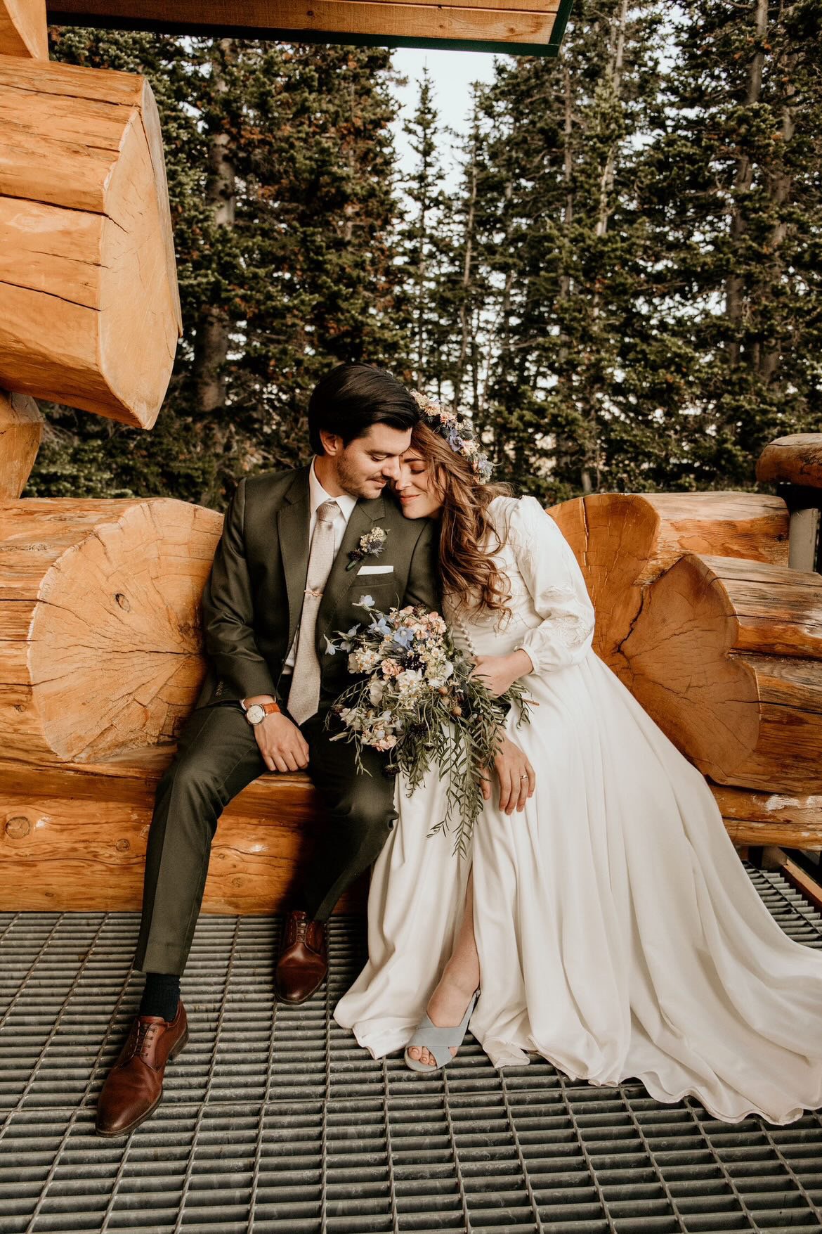 A bride and groom sitting closely, touching foreheads, outdoors with tall pine trees in the background, wooden logs behind and a metal grate underfoot. The bride is wearing a long white dress with a floral headband and holding a bouquet. The groom is
