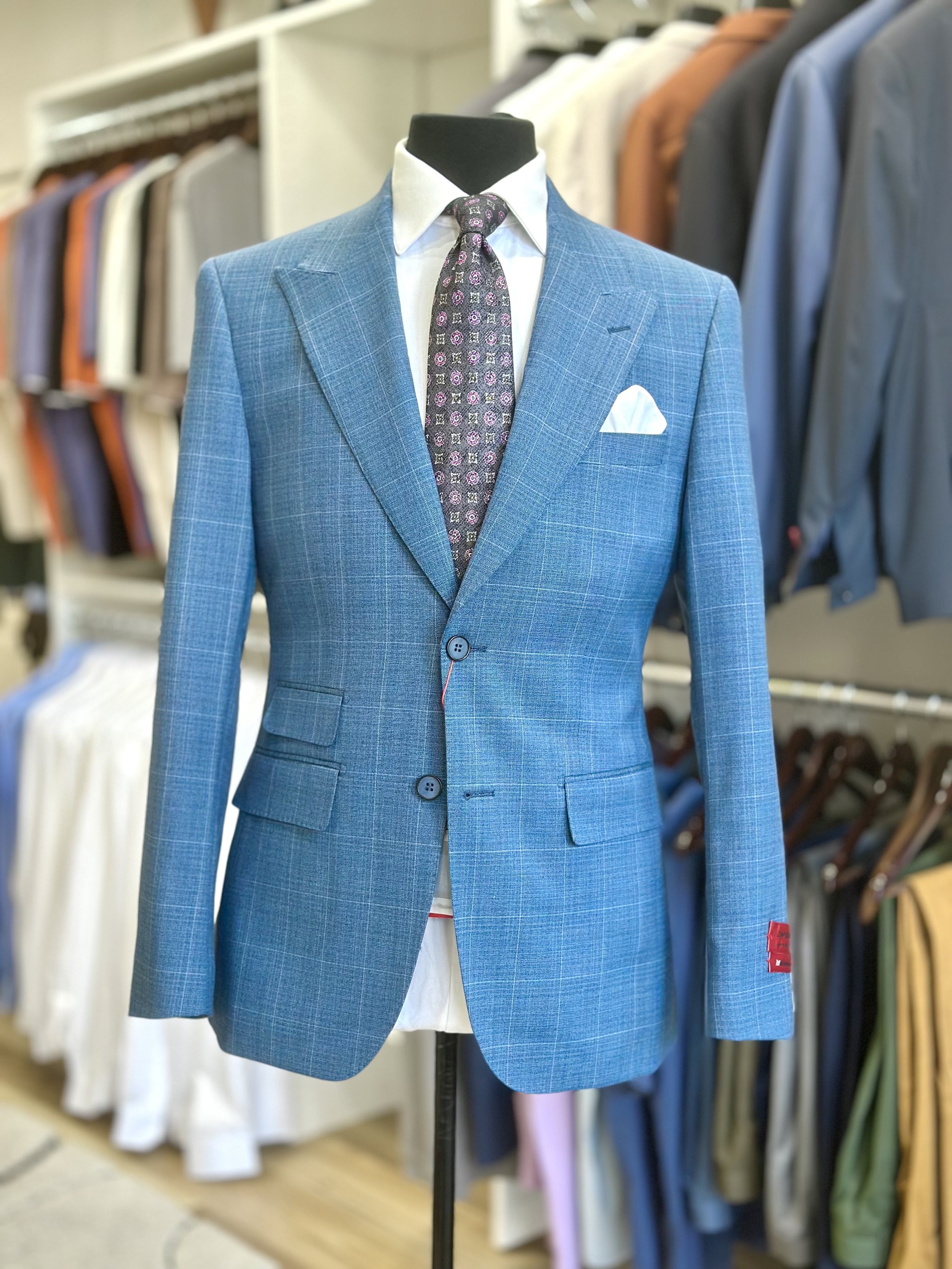 A mannequin dressed in a blue plaid suit jacket, white shirt, and a dark patterned tie, with a white pocket square, displayed in a clothing store with various shirts and suits on racks in the background.