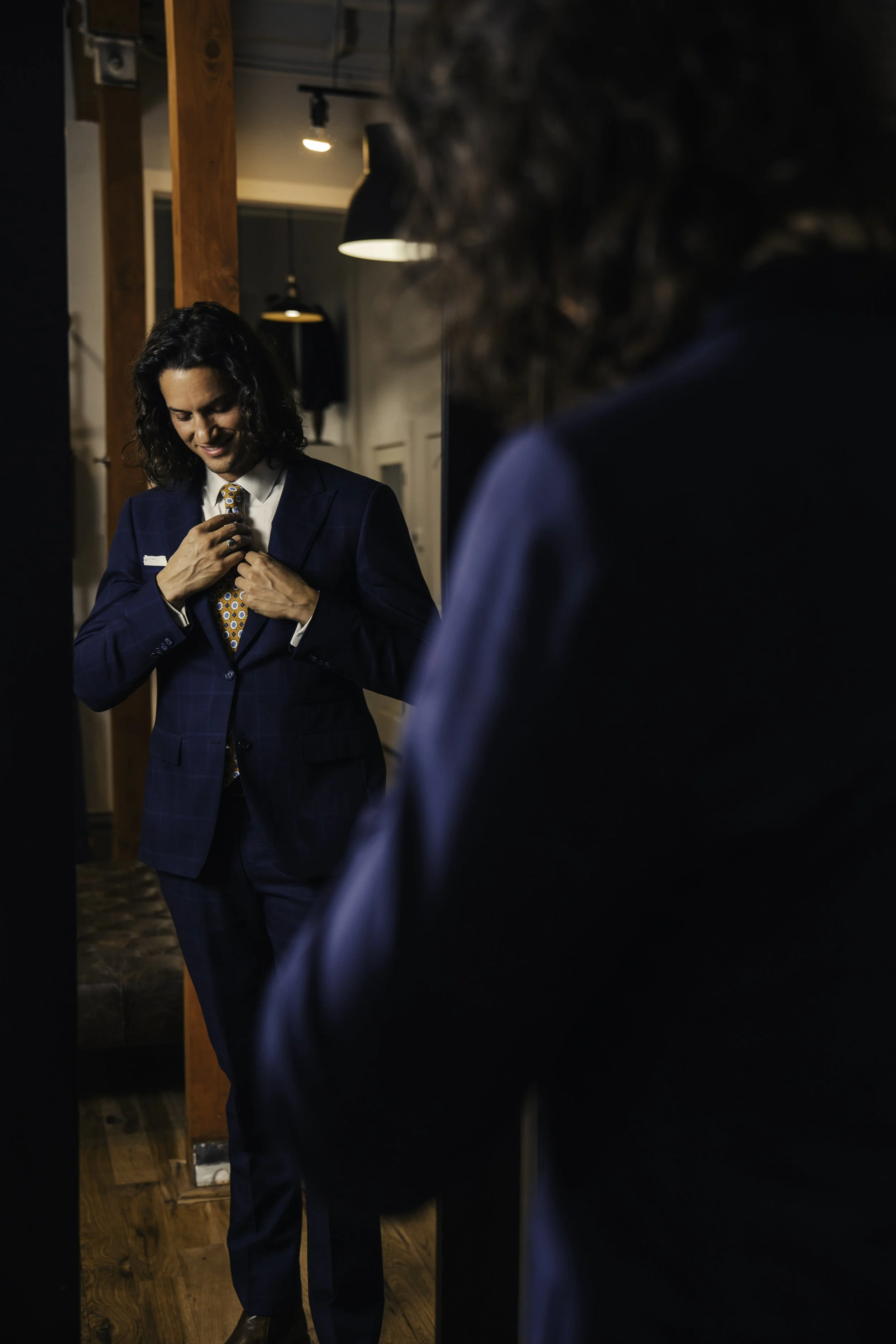 A man in a blue suit adjusting his patterned tie, smiling while looking at himself in a mirror. The setting appears to be indoors with warm lighting.