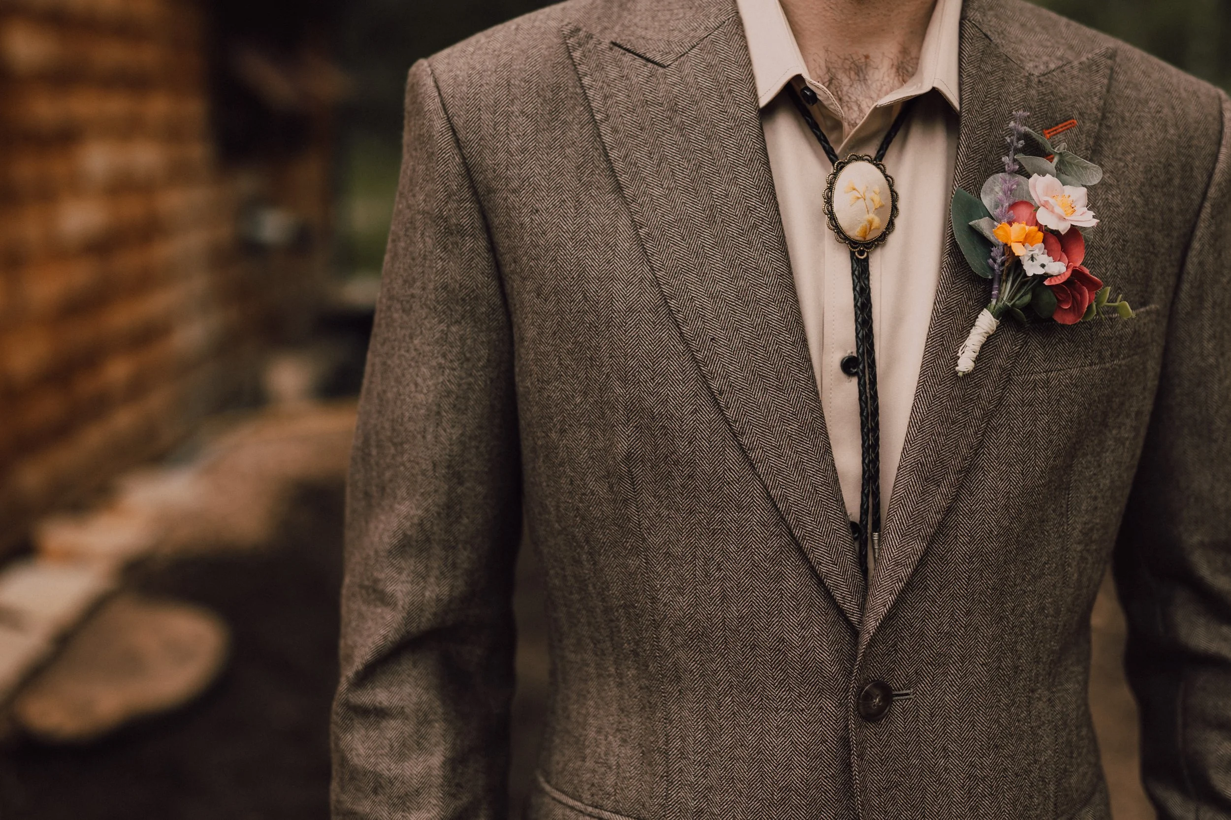 A man wearing a brown herringbone suit with a floral boutonniere on the lapel and a bolo tie with yellow flowers. He is dressed in a beige shirt, seen from the shoulders down.