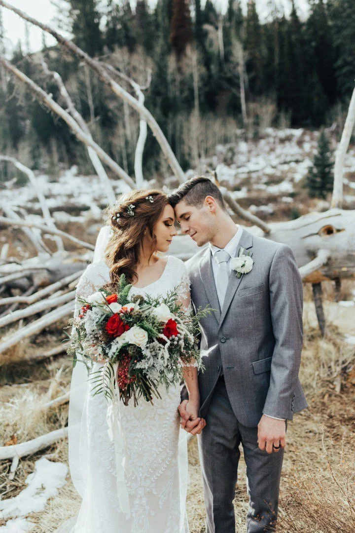 A bride and groom holding hands and touching foreheads outdoors in a forest with fallen logs, some snow, and trees in the background.