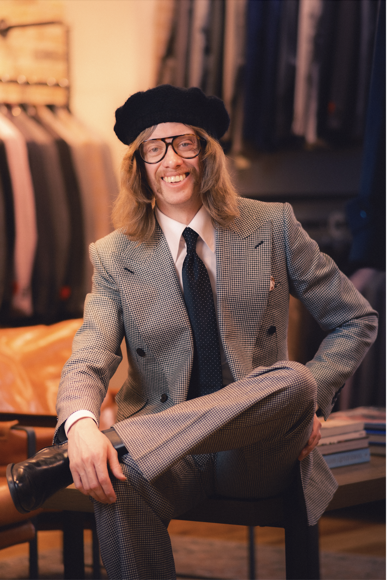 A smiling man with long hair, glasses, and a mustache, wearing a black beret, a checkered suit, a white shirt, and a black polka-dot tie, is sitting on a table inside a room with clothing racks in the background.