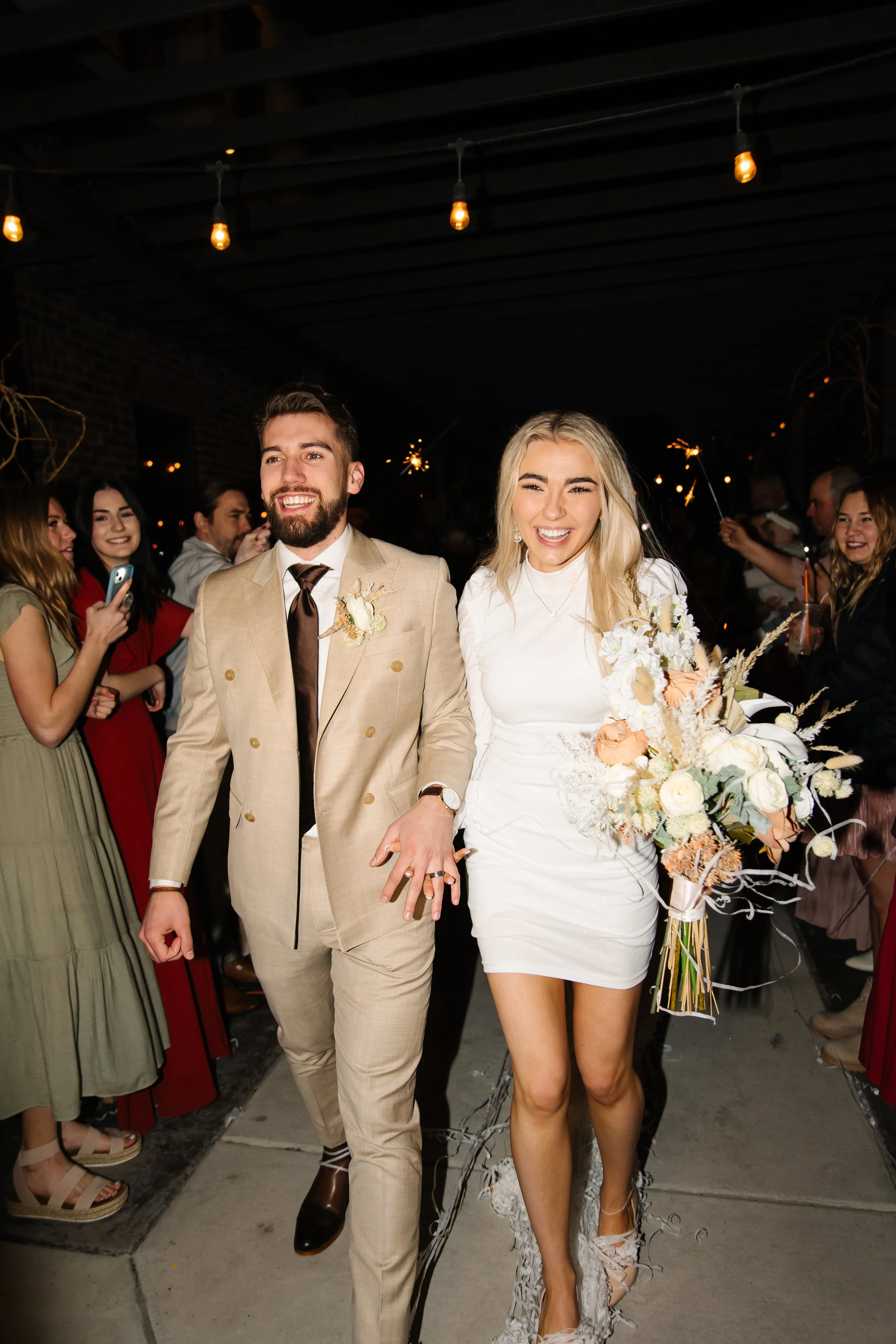 A bride and groom holding hands and smiling at their wedding celebration, surrounded by guests holding sparklers at night.