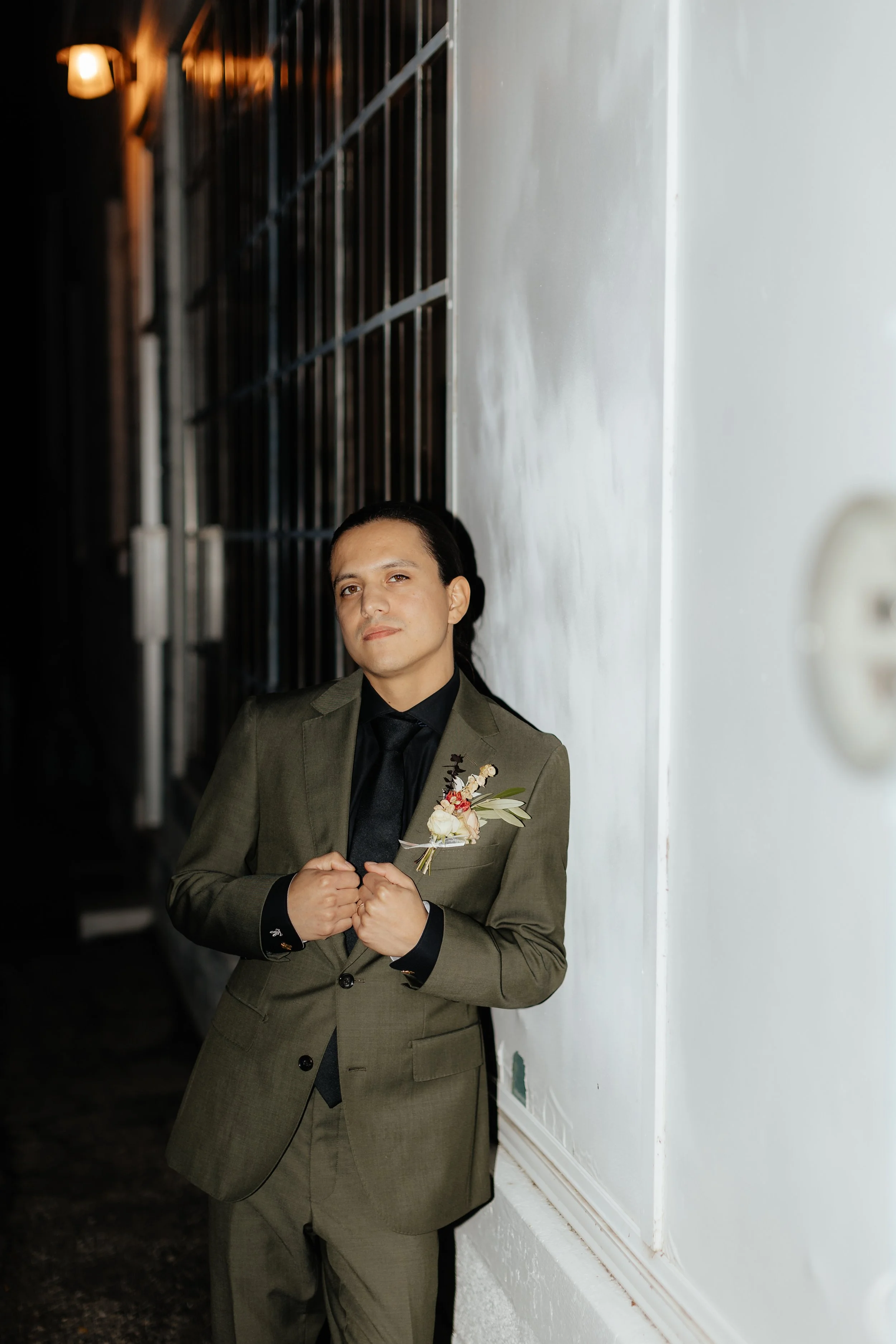 A man in a gray suit holding a small bouquet of flowers stands against a white wall, with a black background.