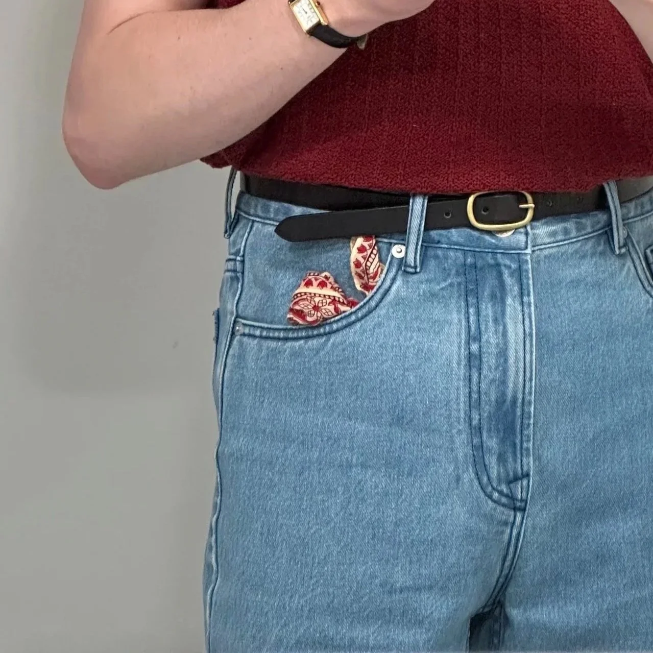 Person wearing light blue jeans with a red patterned bandana partially visible in the coin pocket, a black belt, and a red shirt. The person's wrist has a gold watch and a black wristband.