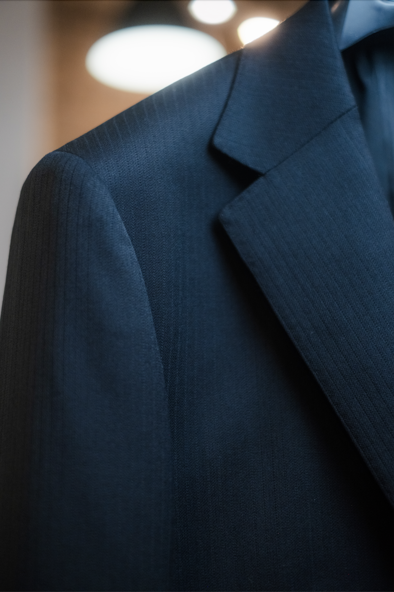 Close-up of a black tailored suit jacket with a notched lapel, featuring textured fabric with subtle stripes, against a blurred background with circular light orbs.