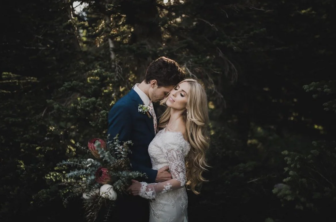 A couple in wedding attire standing close together in a forest, with the groom in a blue suit holding a bouquet and the bride in a white lace wedding dress with long, wavy blonde hair, surrounded by dark green trees.