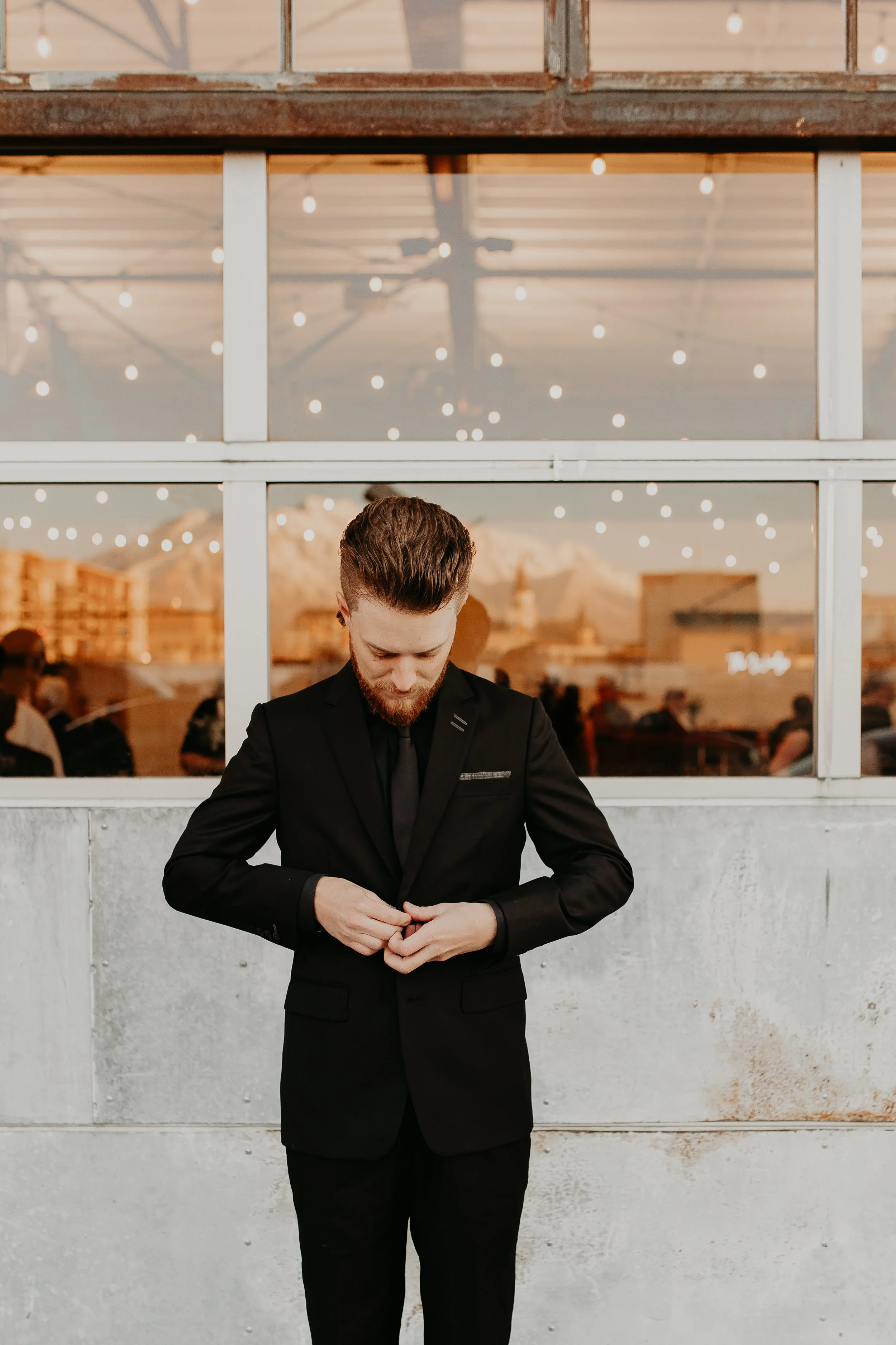 A man with brown hair and a beard wearing a black suit is adjusting his cuffs in front of a window, with a view of a city skyline and mountains at sunset. There are string lights hanging in the background.
