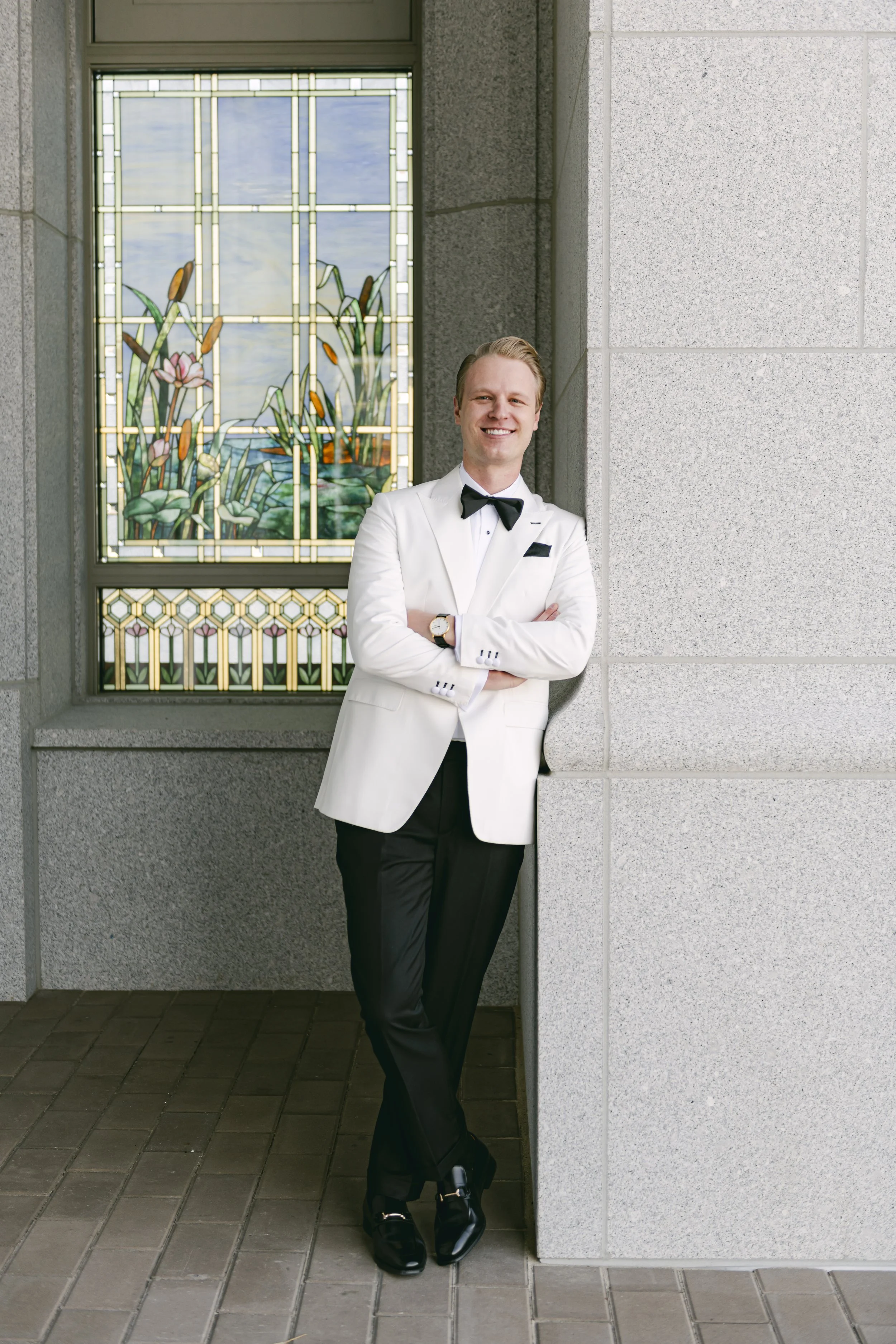 A smiling man in a tuxedo with a white jacket and black bow tie leaning against a wall with his arms crossed outside a building with a stained glass window.