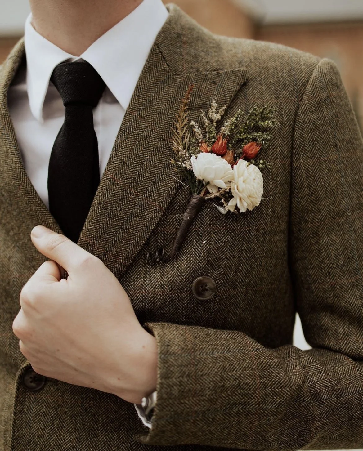 Close-up of a man wearing a brown herringbone suit with a white shirt and black tie, and a boutonniere with white and red flowers.