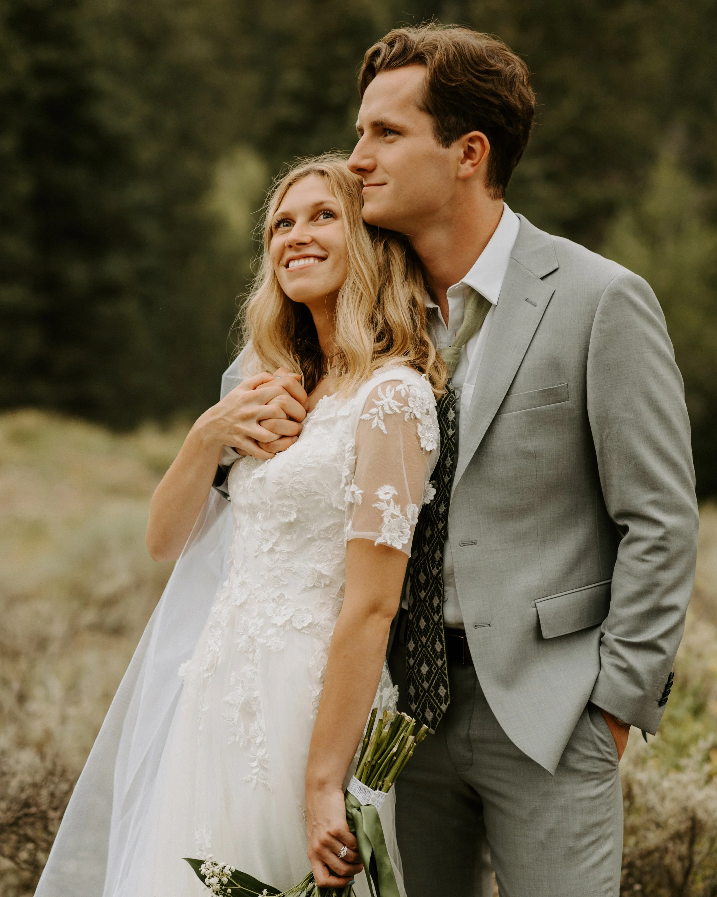 A happy couple on their wedding day standing outside in a natural setting, holding hands, with the groom dressed in a light gray suit and the bride in a white lace wedding gown.