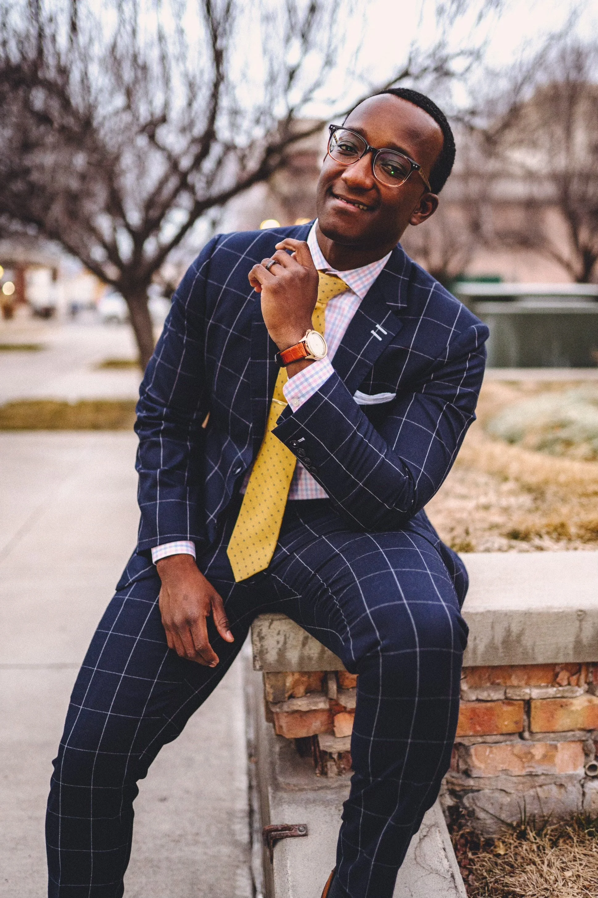 A man dressed in a dark blue checkered suit, yellow tie, and glasses, sitting on a brick ledge outside near bare trees, smiling and adjusting his collar.