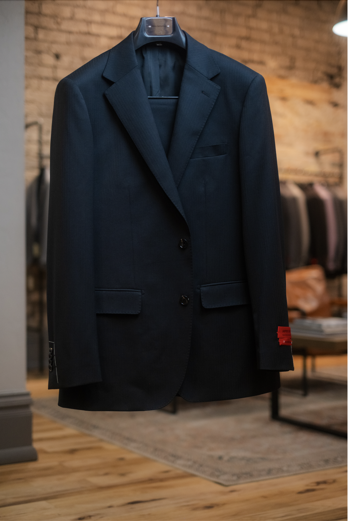 A black suit jacket hanging on a hanger in a clothing store with a brick wall background.