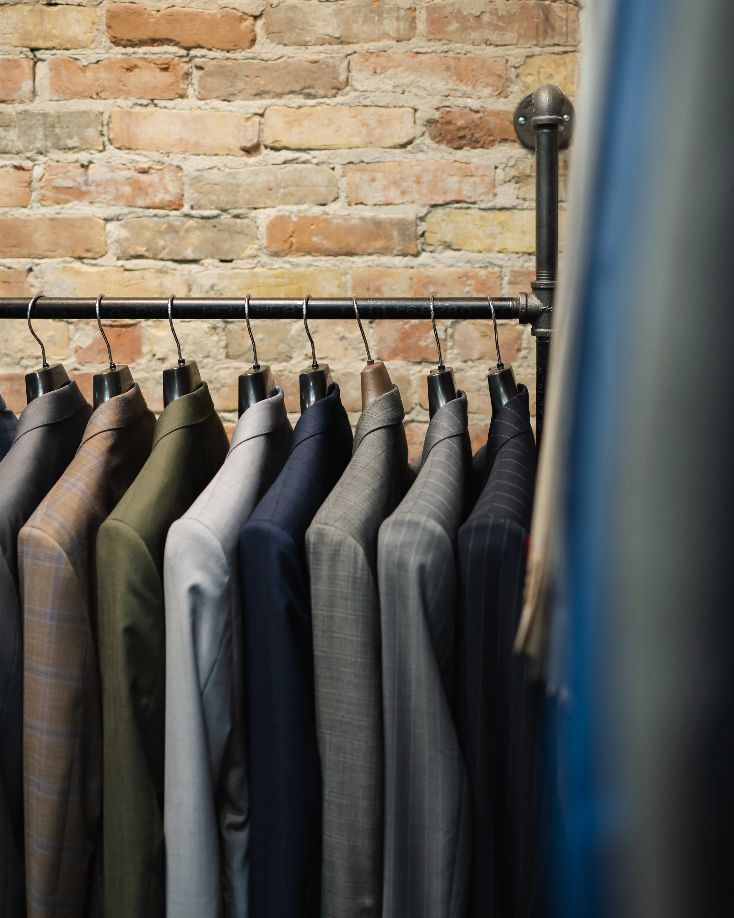 A clothing rack with men's tailored suits in various shades of gray, brown, and blue, hanging against an exposed brick wall.