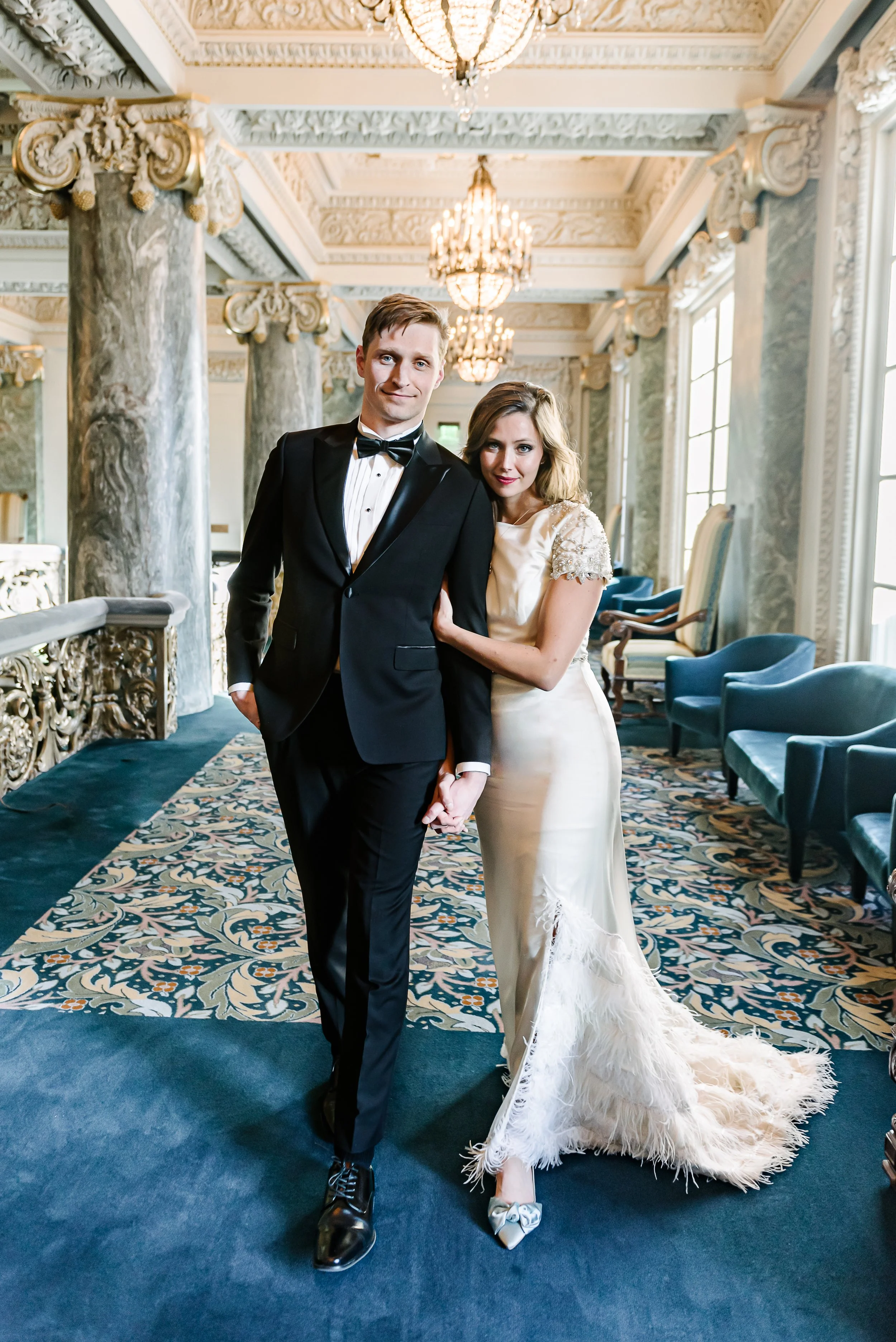 A couple dressed in formal wedding attire standing in an ornate, elegant room with chandeliers, marble columns, large windows, and a patterned carpet.