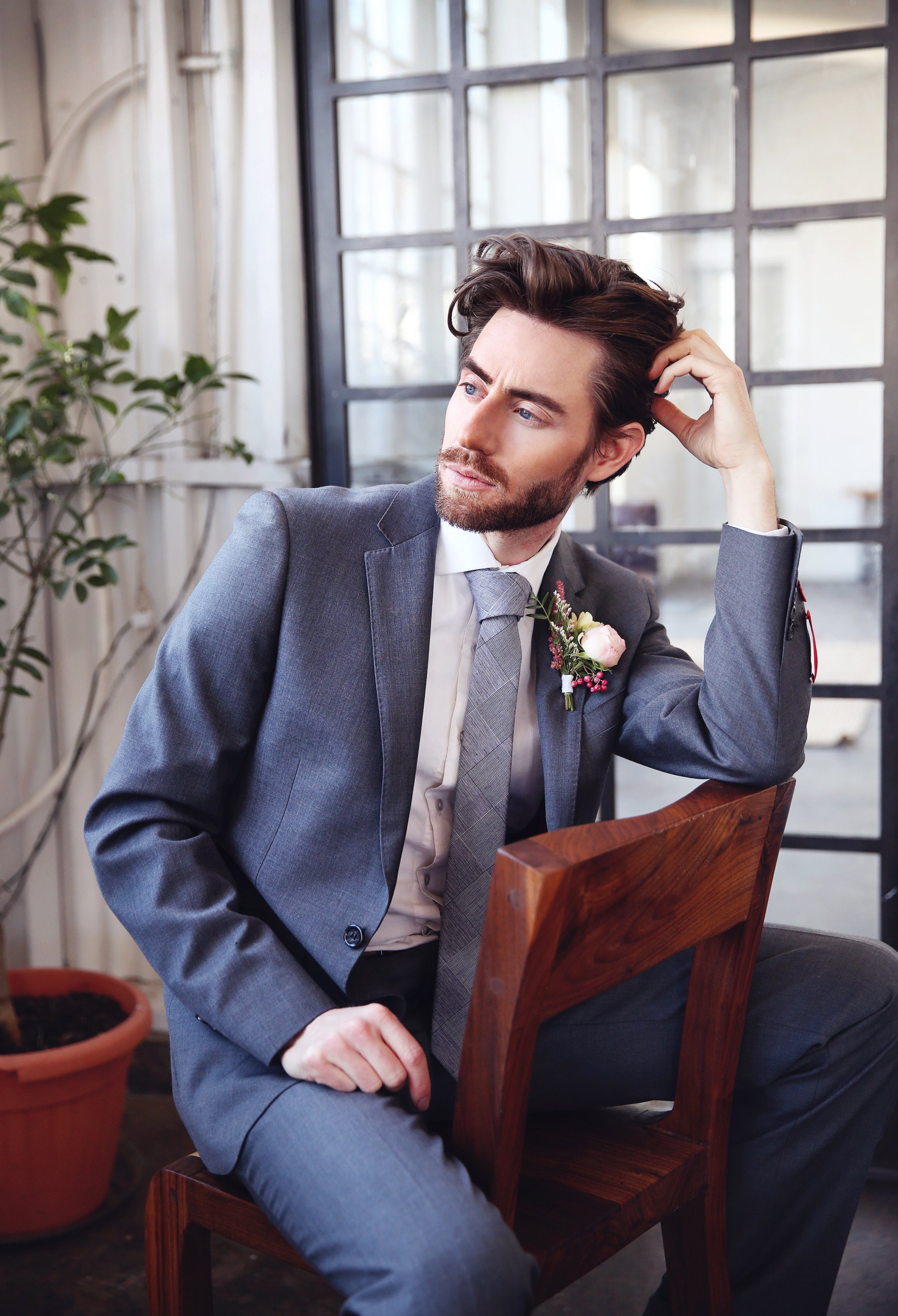 A man with dark hair and a beard, dressed in a gray suit with a boutonniere, sits on a wooden chair indoors. He is resting his head against his hand and looking pensively to the side. The background features large windows and some indoor plants.