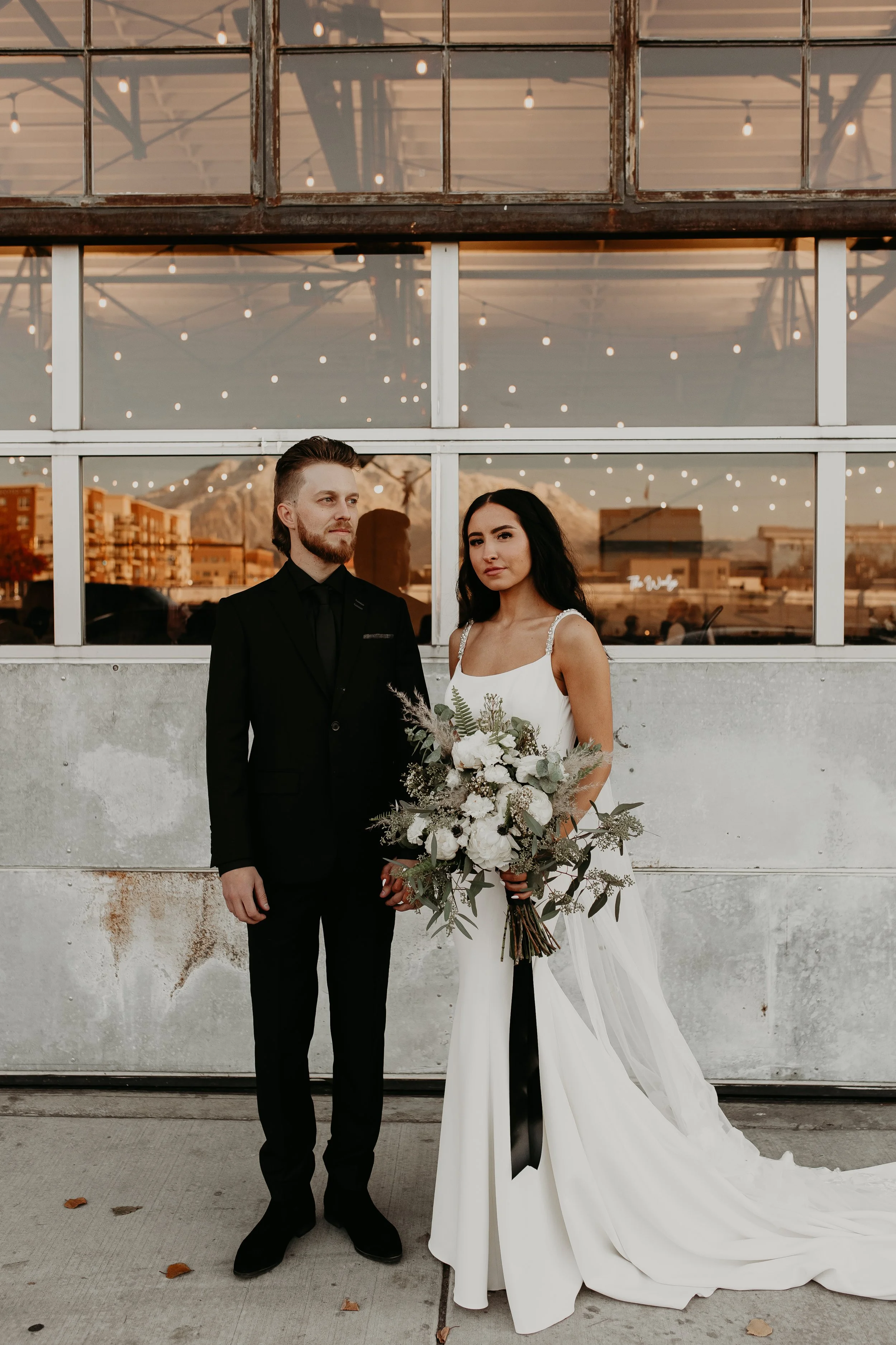 A couple dressed in wedding attire standing outside in front of a building with large windows and string lights. The man is wearing a black suit and the woman is in a white wedding gown holding a bouquet of white flowers and greenery.