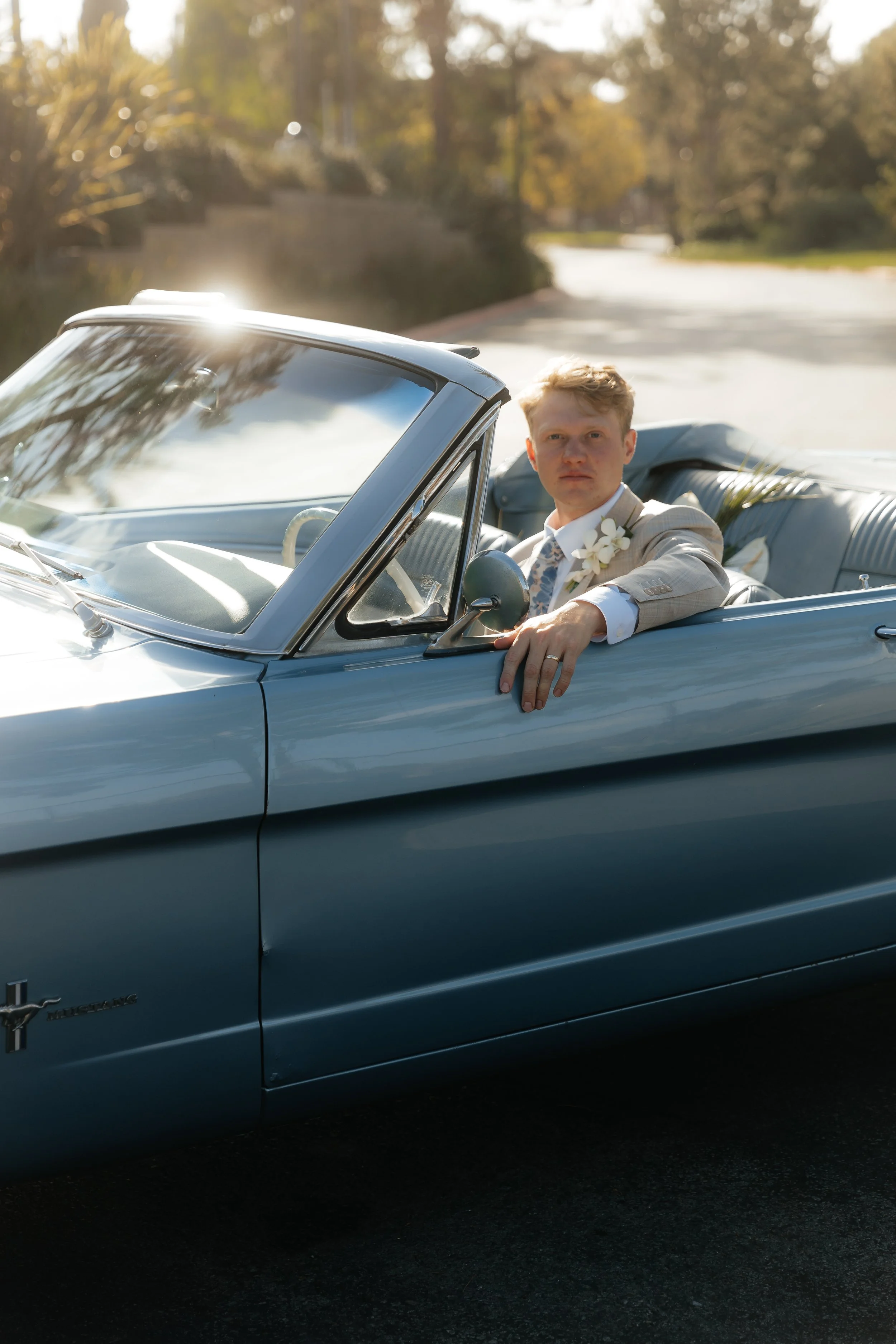 A young man in a light-colored suit sitting in the driver's seat of a vintage blue convertible car, parked on a suburban street with trees and houses in the background.