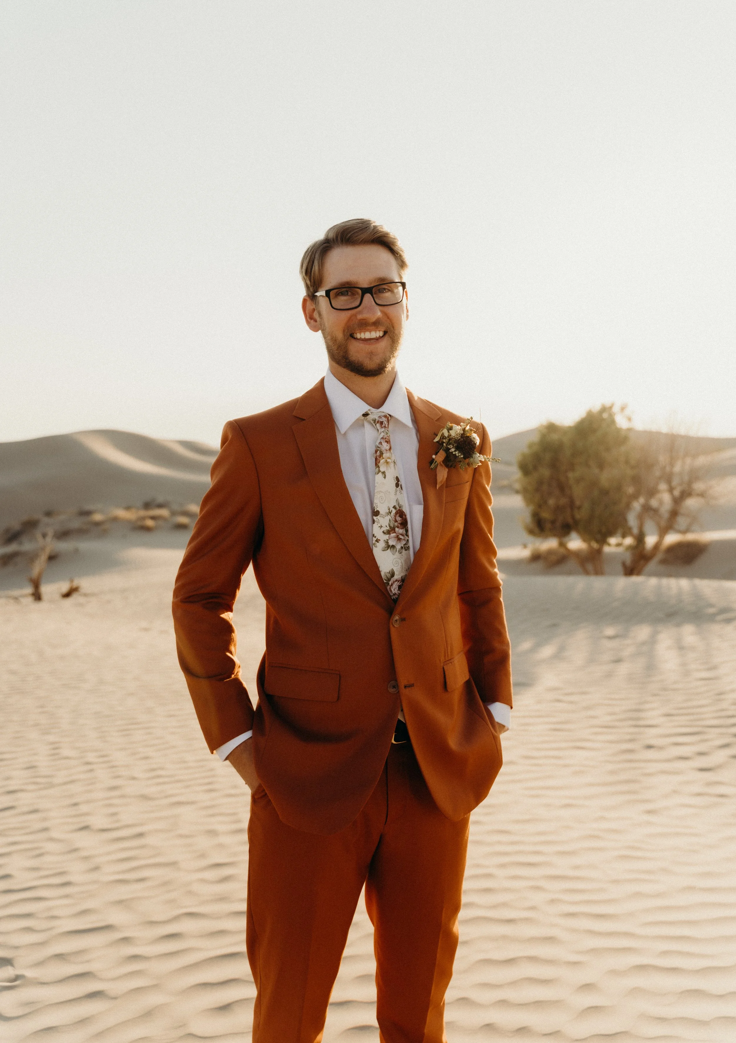 A smiling man in a rust-colored suit, white shirt, floral tie, and glasses stands in a desert landscape with sand dunes and sparse trees during sunset.